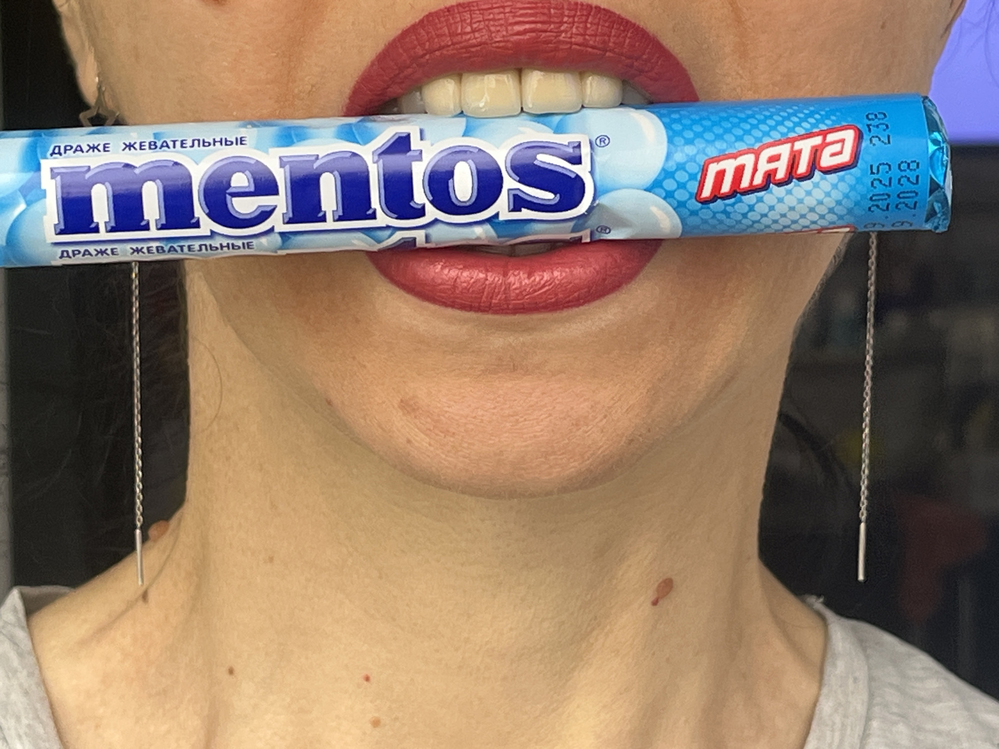 A close-up, medium shot depicts a woman holding a roll of Mentos Mata gum in her mouth. Her lips are painted with a matte red lipstick. The gum wrapper is blue and white, with the word "Mentos" printed in blue. The label also has the word "Mata" in red. There are two silver, threader earrings on either side of the woman's face. The date "3.2025 238 3.2028" is visible in tiny print on the right side of the gum wrapper. The woman is wearing a gray t-shirt. There are various freckles/moles on the woman's face and neck.
This product is branded as being sold in Krasnodar, Russia.