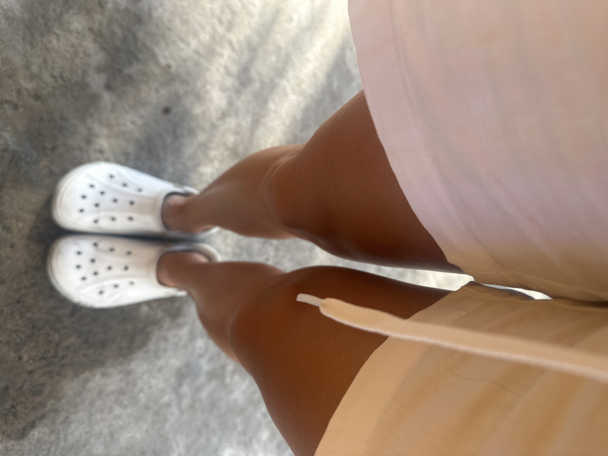 A low-angle, eye-level shot captures a person standing on a concrete surface. The person is wearing white slip-on shoes with circular perforations and a white or light-colored dress or shorts. The legs are tanned. The background is a gray concrete surface, possibly a sidewalk or floor. The location is Bac Nha Trang Ward, Vietnam.