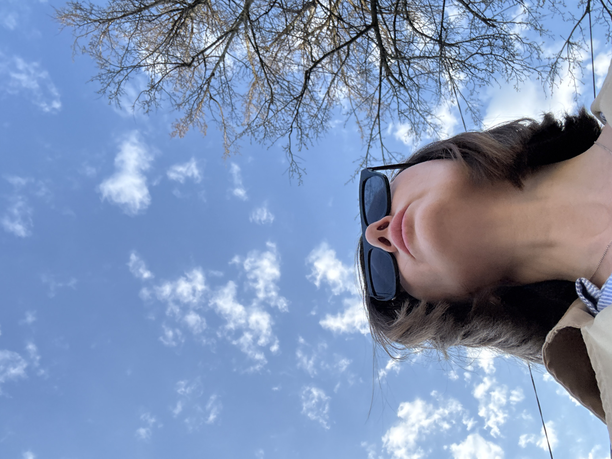The image is a low-angle shot looking upwards towards a blue sky with scattered white clouds and bare tree branches at the top. A person is partially visible in the foreground, wearing sunglasses. The setting appears to be outdoors during the daytime.
It is not possible to specifically confirm the location as Chaikovsky, Russia.