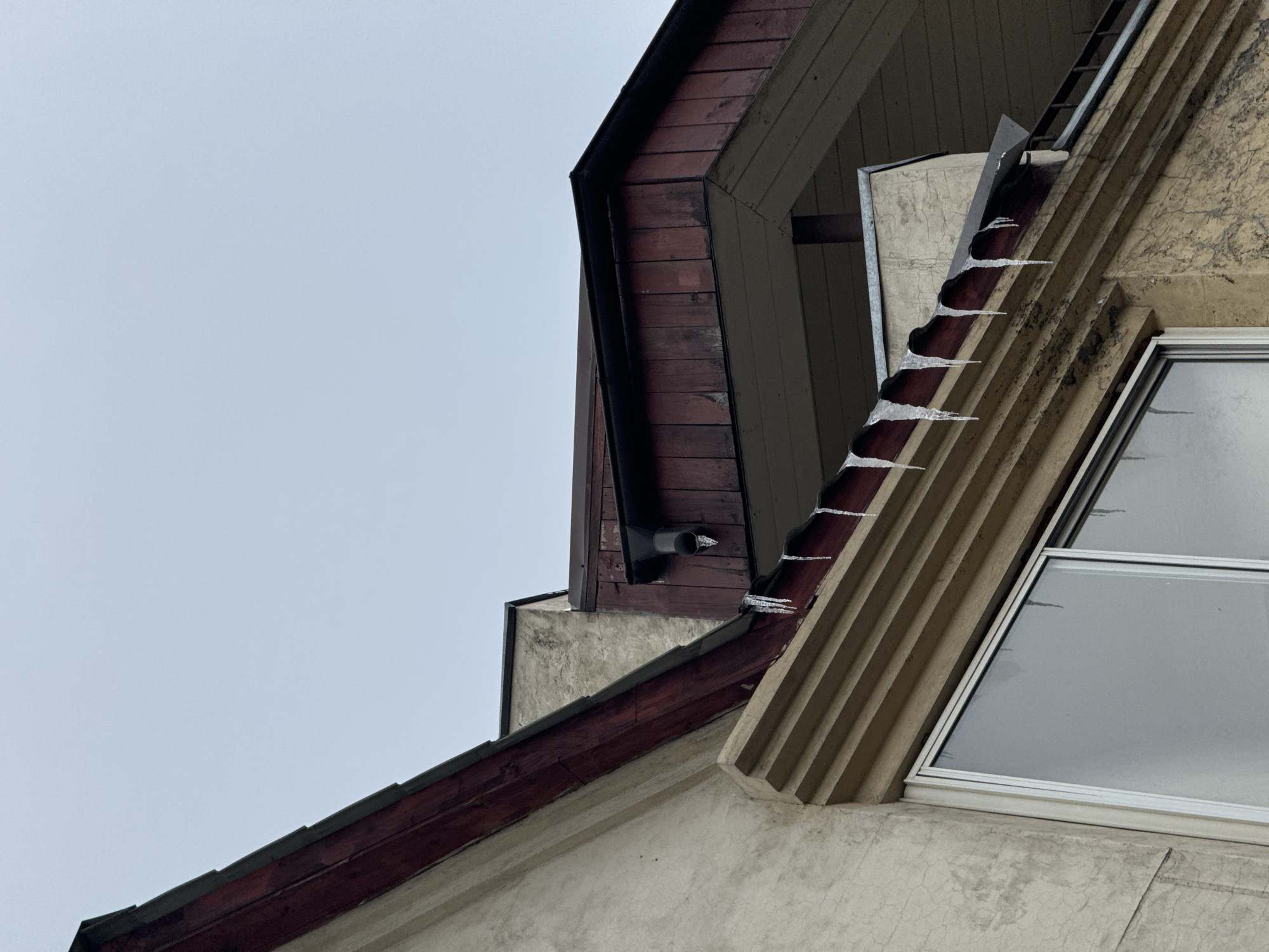 The image depicts a building facade in Krasnoyarsk, Russia, with a complex roofline, observed during cold weather conditions, indicated by the presence of icicles. The architecture incorporates various materials and textures, including painted stucco, dark-stained wood, and glass windows. The facade features a window with a white frame, set adjacent to fluted, vertical detailing. A dark-colored drainage pipe protrudes from a wooden section of the roof. The sky is overcast.