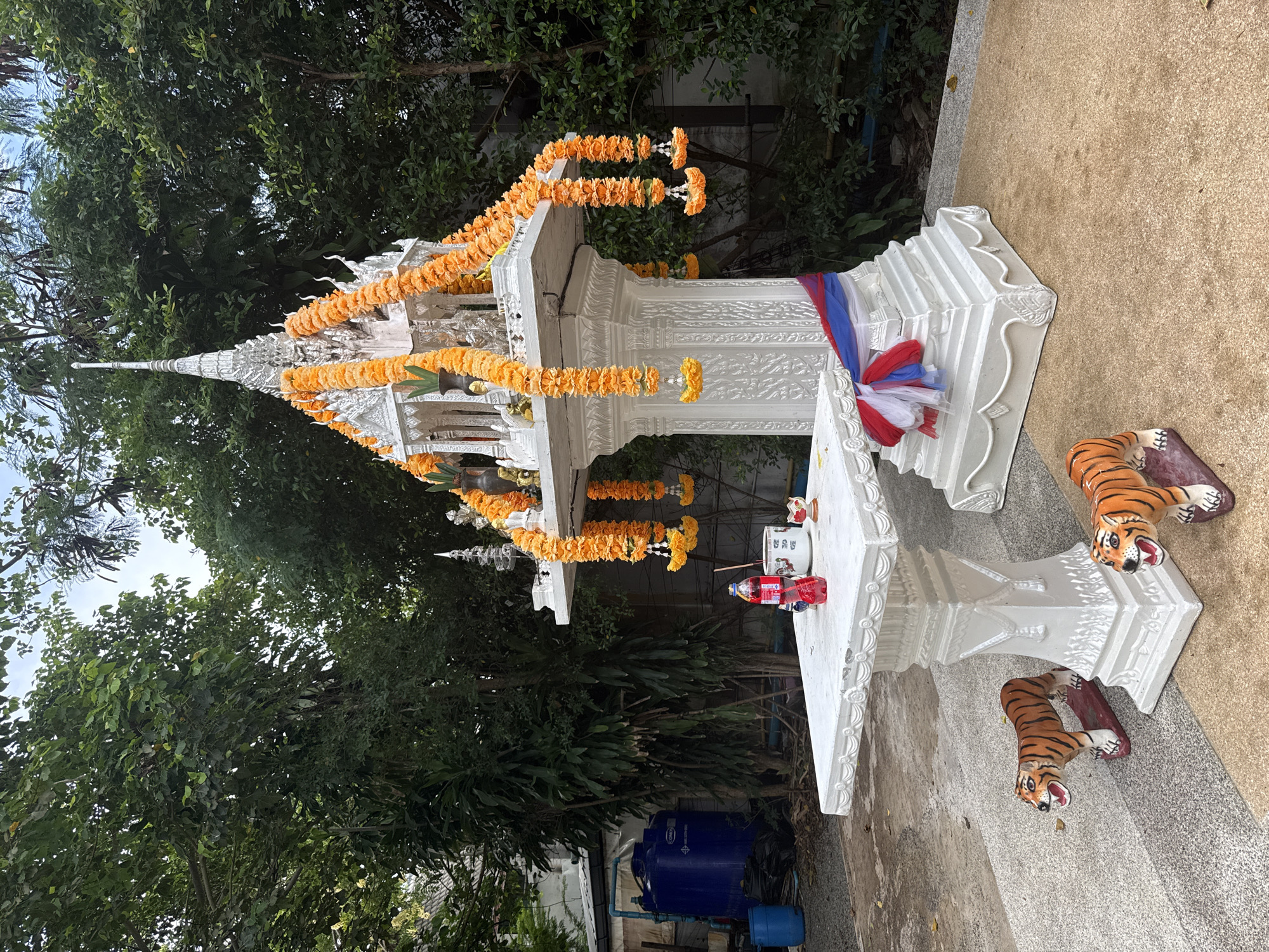 The image shows a white spirit house decorated with orange garlands, situated in Rawai, Thailand. In front of the spirit house is a white table with a red bottle of Coca-Cola, a cup, and a few other small offerings. Two tiger statues stand guard on either side of the table. The spirit house is adorned with intricate carvings and has a pointed roof. The area is surrounded by lush green trees, suggesting a tropical environment. In the background, a large blue water tank is visible. The ground is a mixture of concrete and small pebbles.