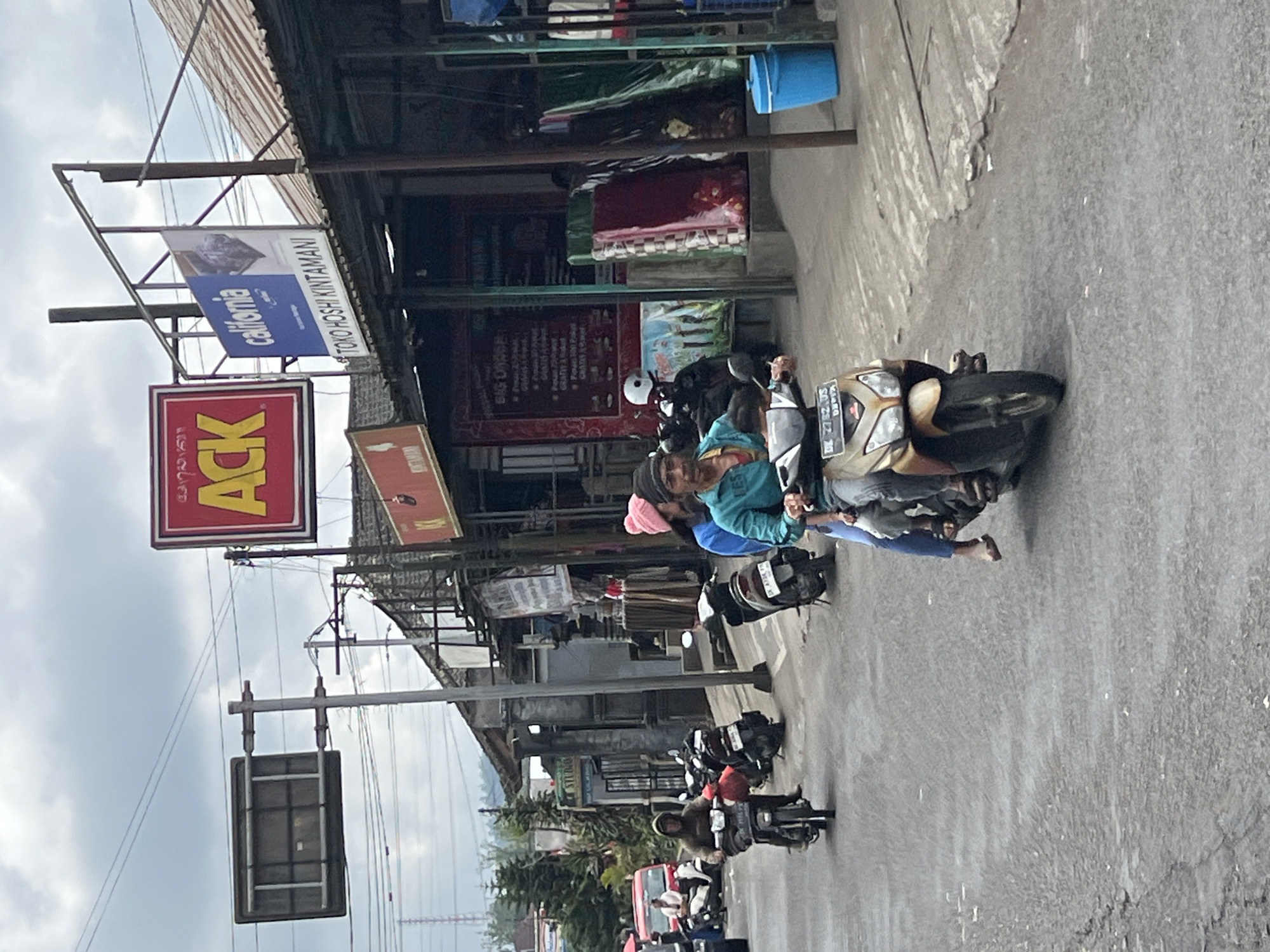 The image depicts a street scene in Kintamani, Indonesia, under a cloudy sky. In the foreground, two people ride a gold-colored scooter. The driver wears a blue-green shirt and a dark beanie, while the passenger, a woman, wears a blue shirt and a pink beanie. They are on a paved road with other scooters and a red van visible further down. Shops line the right side of the street, with signs indicating "ACK" and "California TOKO HOSHI KINTAMANI". Several scooters are parked along the roadside, and some products, possibly mattresses, are displayed outside one of the shops. The overall atmosphere suggests a typical day in a bustling Indonesian town.