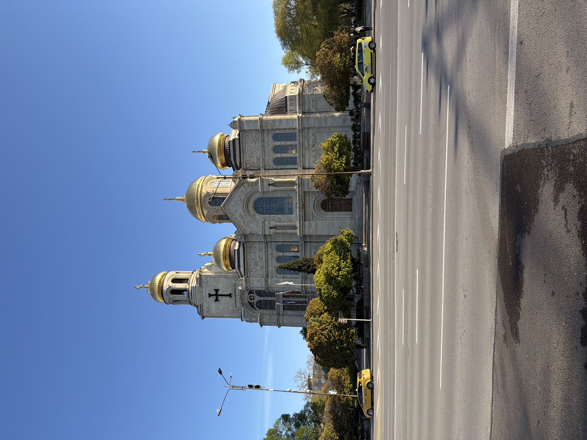 The image shows the Dormition of the Mother of God Cathedral in Varna, Bulgaria. The cathedral is a large, ornate building with multiple golden domes and a white facade. It is positioned adjacent to a wide, multi-lane street marked with white lines. Several trees are planted along the side of the cathedral and the street. Two yellow vehicles are parked on the side of the road. A light post is visible near the bottom of the frame. The sky is clear and blue.