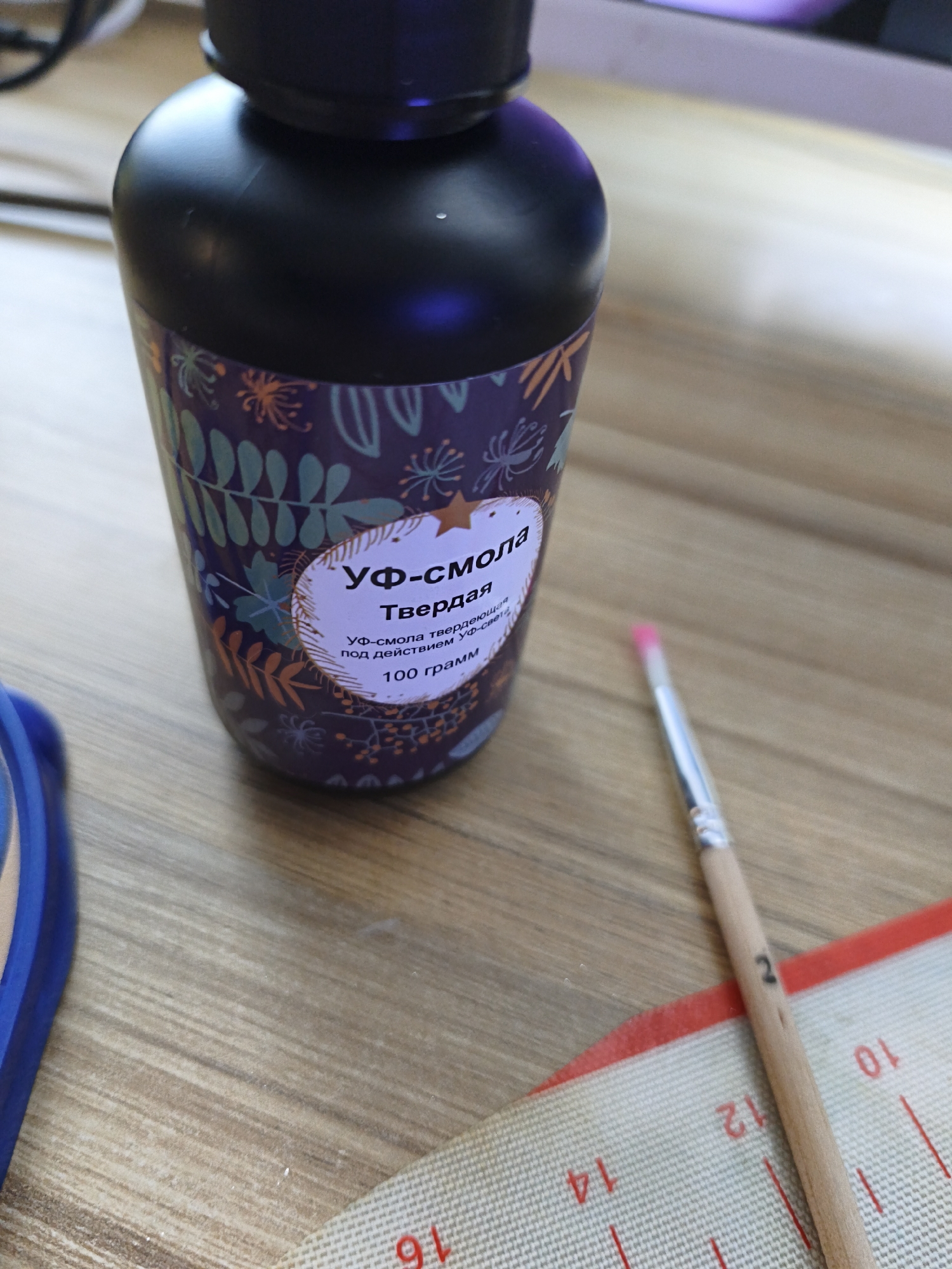 The image displays a close-up of a dark purple bottle of UV resin and a paintbrush on a light brown wooden surface. A silicone mat with numerical markings is partially visible in the foreground, with a blue object at the far left.
The bottle, centrally positioned, has a black cap and a colorful label featuring stylized leaves and branches in green, orange, and blue hues on a dark purple background. An oval section on the label contains Cyrillic text. The text reads "УФ-смола" (UV resin), "Твердая" (Hard), followed by "УФ-смола твердеющая под действием УФ-света" (UV resin hardening under the action of UV light), and "100 грамм" (100 grams).
To the right of the bottle, a paintbrush with a wooden handle and a vibrant pink bristle tip is laid horizontally. The number "2" is subtly marked in black on the brush handle. In the lower right of the frame, a textured white silicone mat with a red border is visible. Red lines and numbers are printed on the mat; discernible numbers include "10", "12", "14", and "16", some appearing upright and others inverted, indicating a measuring scale or grid. The scene appears to be an indoor setting, possibly a crafting area, under soft illumination.
