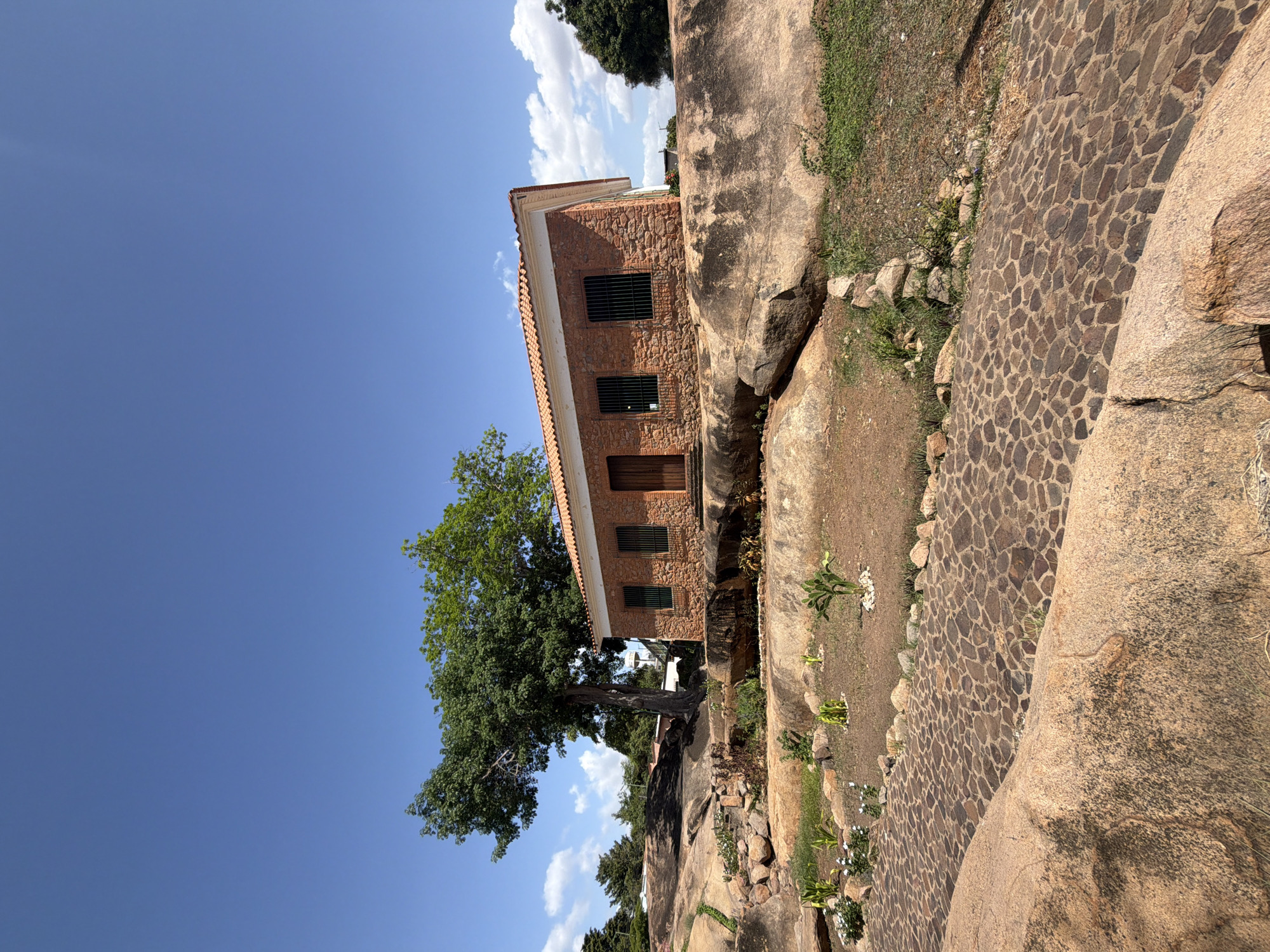 A sideways, wide shot captures a two-story, stone brick building with a red tile roof in Ciudad Bolivar, Venezuela. The building features barred windows and a wooden door. A large, leafy tree stands beside the building, partially obscuring its facade. The structure is built atop a large rock formation, and a stone-paved walkway runs parallel to the building. The walkway is bordered by additional rocks and sparse vegetation. Above, a clear blue sky is dotted with a few white clouds.