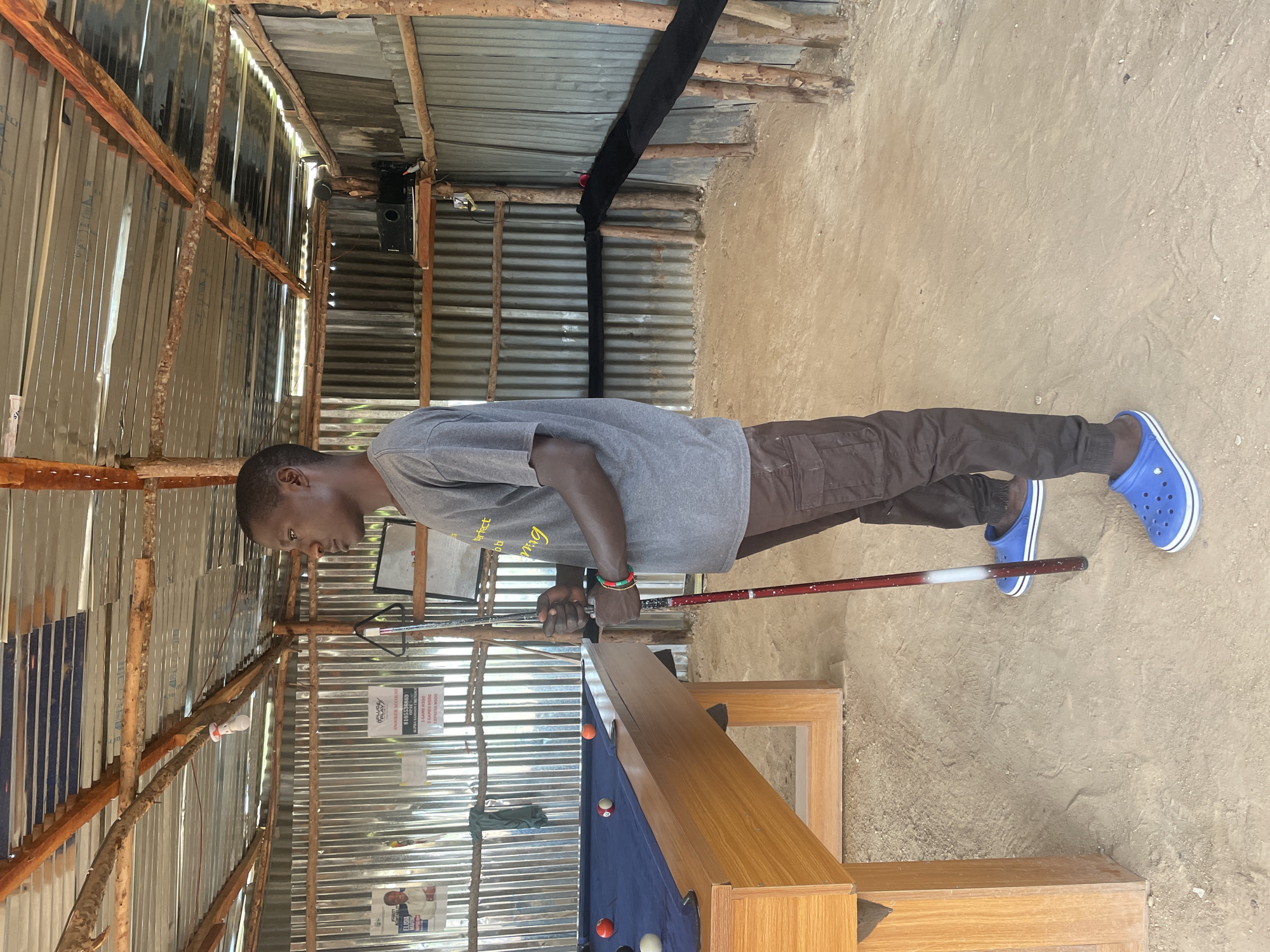 In Durumi, Nigeria, a young man stands beside a pool table in an outdoor, open-air establishment. The structure features a corrugated metal roof supported by wooden beams and posts, with corrugated metal walls forming the sides. The ground is composed of packed dirt. The man, wearing a grey t-shirt, brown cargo pants, and blue Crocs, holds a red and white pool cue. The pool table, covered in blue felt, has several balls scattered across it. In the background, posters are visible on the corrugated metal wall, and a projector is mounted on a shelf. A black cloth barrier is set up behind the pool table. The lighting suggests it is daytime.