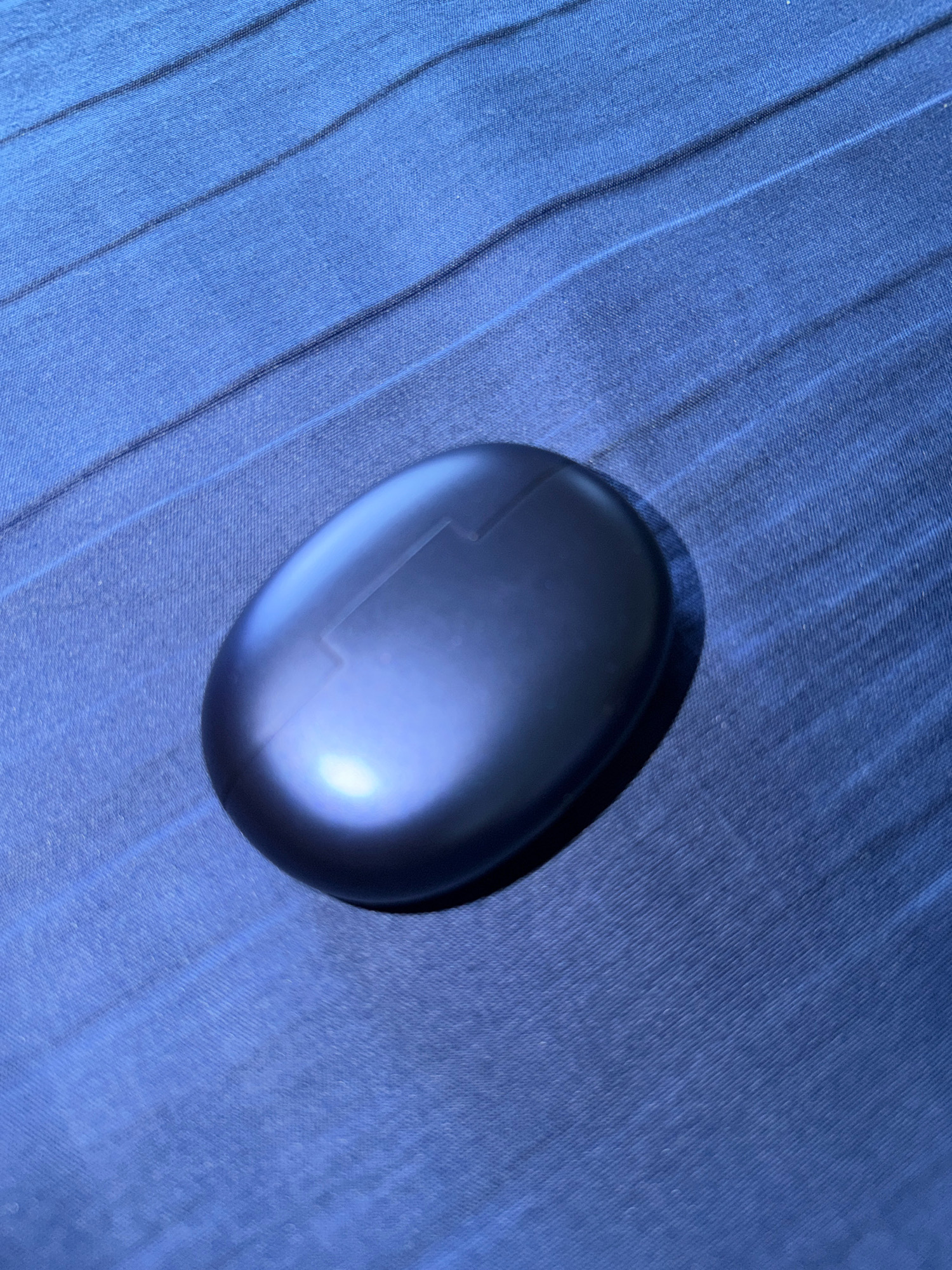 A dark blue oval-shaped charging case for wireless earbuds rests on a blue fabric surface. The fabric has a textured, ribbed pattern. The case has a subtle seam or design element visible on its top surface. The lighting creates a bright reflection on the case, and a soft shadow to its right. The scene is not in Kriparampur, India.