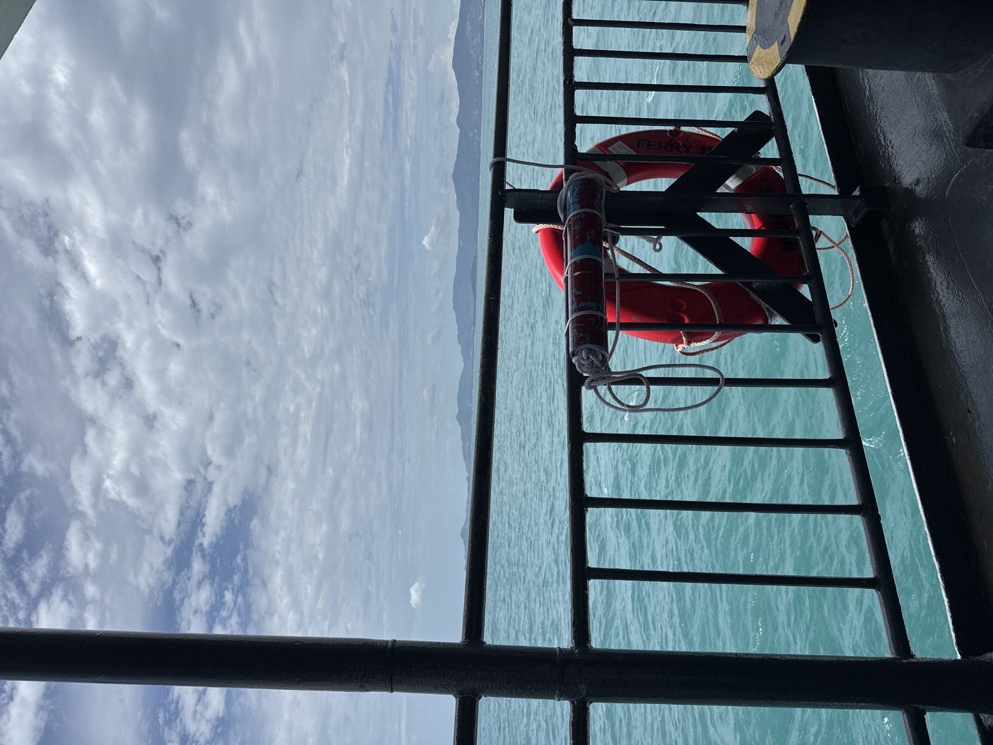 A life preserver hangs on a black metal railing on the deck of a ferry in the Ko Pha-ngan District of Thailand. The sea is a vibrant turquoise and appears calm, with gentle waves. In the distance, a hazy, mountainous coastline is visible under a cloudy sky. The railing is positioned in the foreground, partially obscuring the view of the sea and the distant land. A thick, dark pole is also visible on the left side of the frame, indicating the structure of the ferry. The life preserver is red and has some white and blue markings. A rope is attached to it and tied to the railing. The deck of the ferry is wet and appears to be made of dark material.