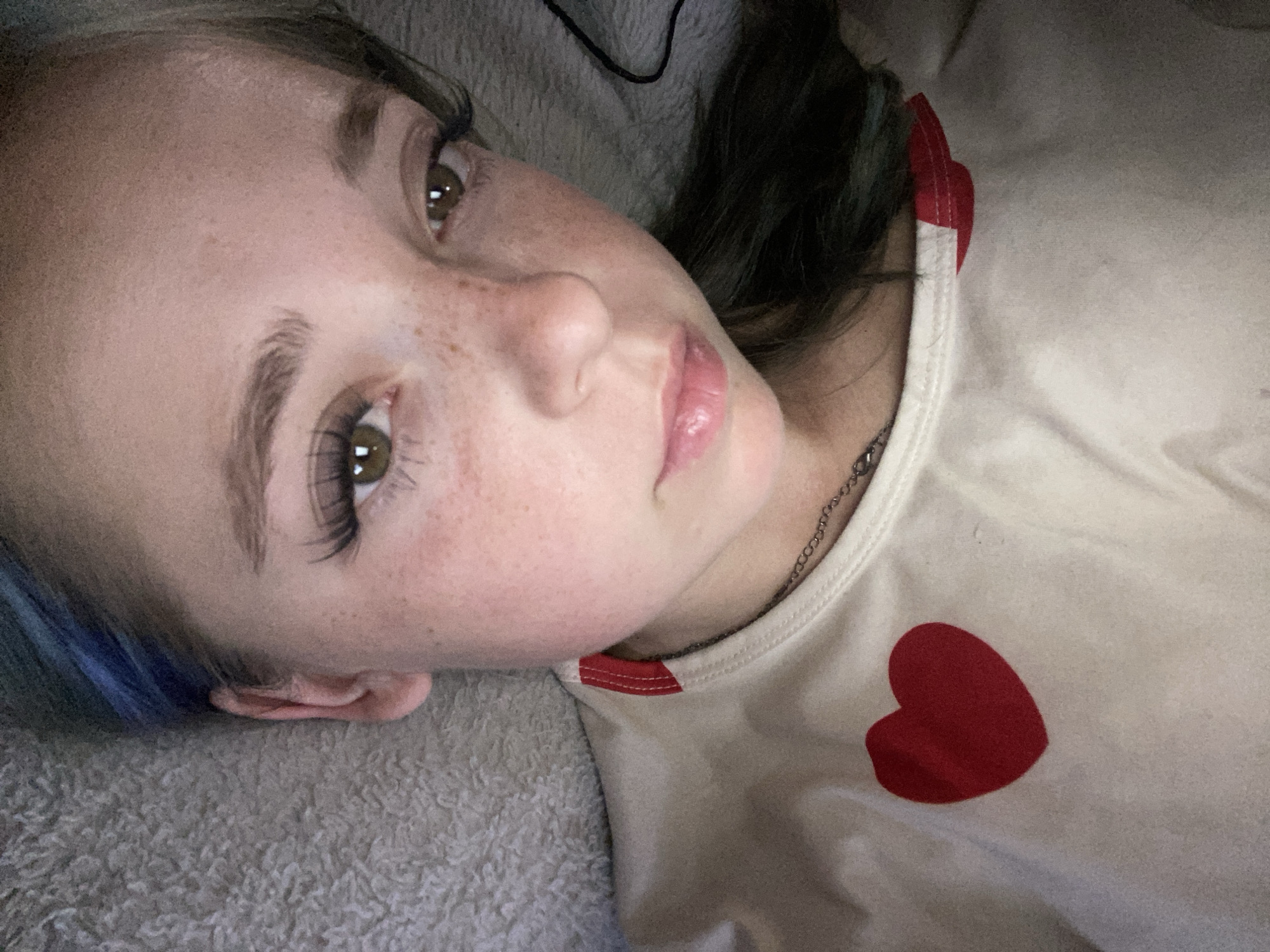 This is a close-up, overhead, indoor photograph of a young person lying down. The person has light skin with freckles, green eyes, and dark hair with blue streaks. They are wearing a light-colored t-shirt with a red heart graphic on the left side of the chest and a red trim around the neckline. The background is a textured, light-colored surface. There are no discernible landmarks or contextual clues that would allow for the identification of a specific city, including Nova Praha, Ukraine.
