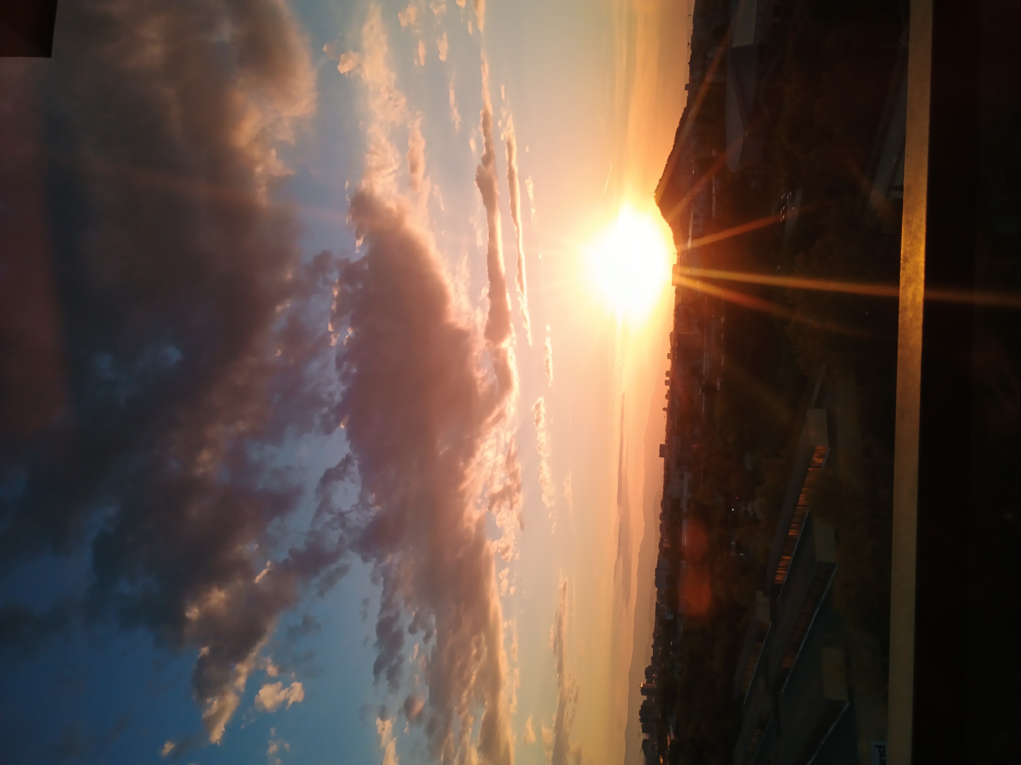 The image captures a sunset over Plovdiv, Bulgaria. The sun is positioned near the horizon, casting intense light and rays across the urban landscape. The sky is filled with clouds in varying shades of blue, grey, and orange, illuminated by the setting sun. The cityscape is visible, featuring buildings and structures against the skyline. A vertical element, likely a window frame or part of a building, is seen on the right.