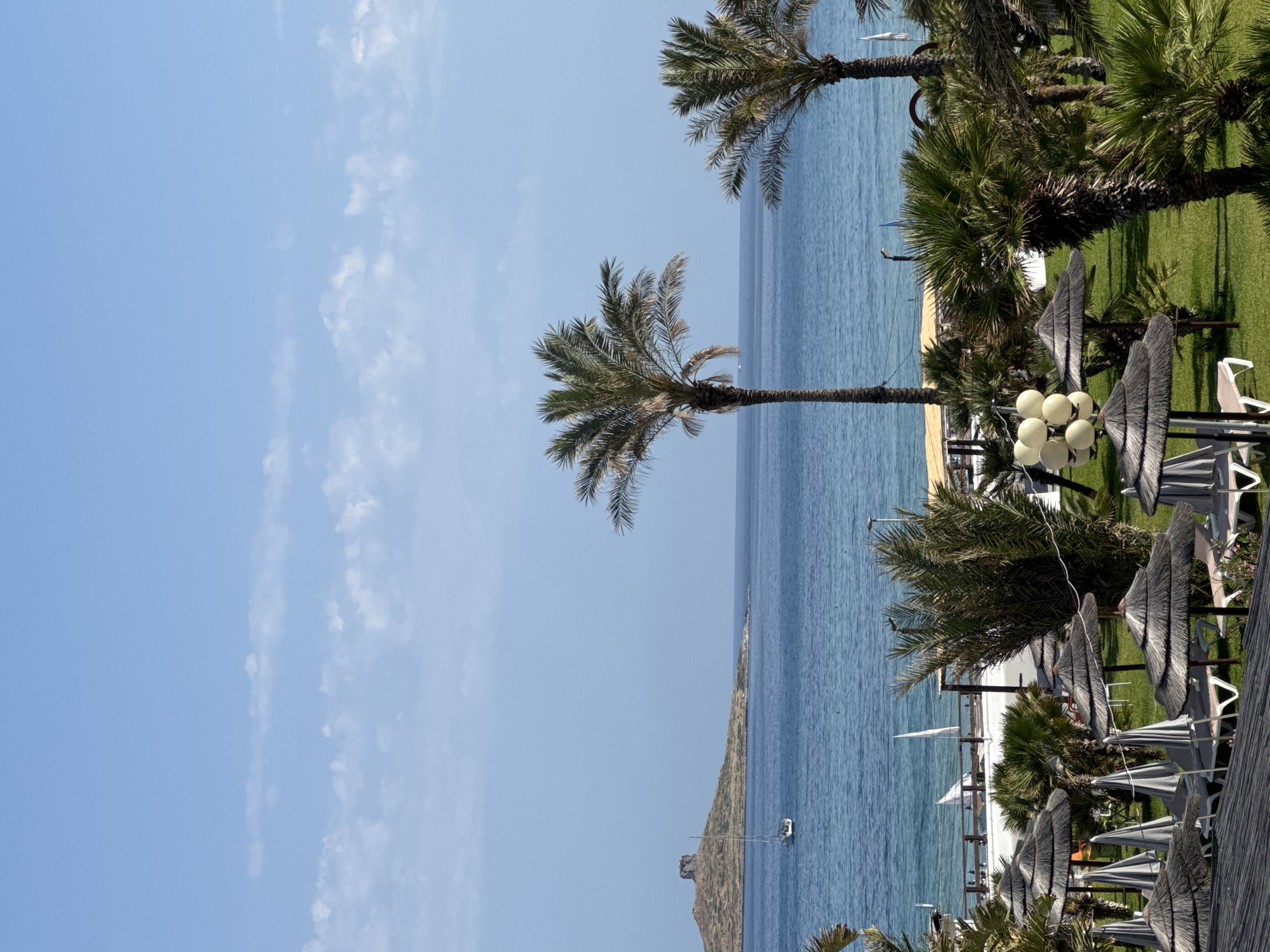 This vibrant image depicts a picturesque coastal resort scene, identified as Isola delle Femmine, Italy. The view is dominated by an expansive, bright blue sky with a few scattered, wispy white clouds, indicative of clear and pleasant weather.
The serene turquoise sea occupies the middle ground, stretching towards a distant, low-lying landmass or island visible on the horizon. A small white boat or yacht can be seen gently floating on the calm waters near this distant land.
Numerous palm trees are prominent throughout the composition, their green fronds contrasting with the blue sky and sea. Some palms stand tall, reaching into the sky, while others line a neatly manicured green lawn that forms the foreground on the right side of the image. Between the green grass and the sea, a narrow strip of sandy beach is visible.
The grassy area is furnished with typical resort amenities, including several thatched beach umbrellas and modern fabric umbrellas, alongside white lounge chairs, suggesting an area dedicated to relaxation and sunbathing. A distinctive lamppost, featuring multiple spherical white light fixtures, is also visible amongst the resort furniture.
The time of day appears to be bright daytime, with ample natural light illuminating the scene, creating a peaceful and inviting atmosphere. There are no people visible in the image, allowing the natural beauty and resort infrastructure to be the primary focus. No visible text is present within the media file.