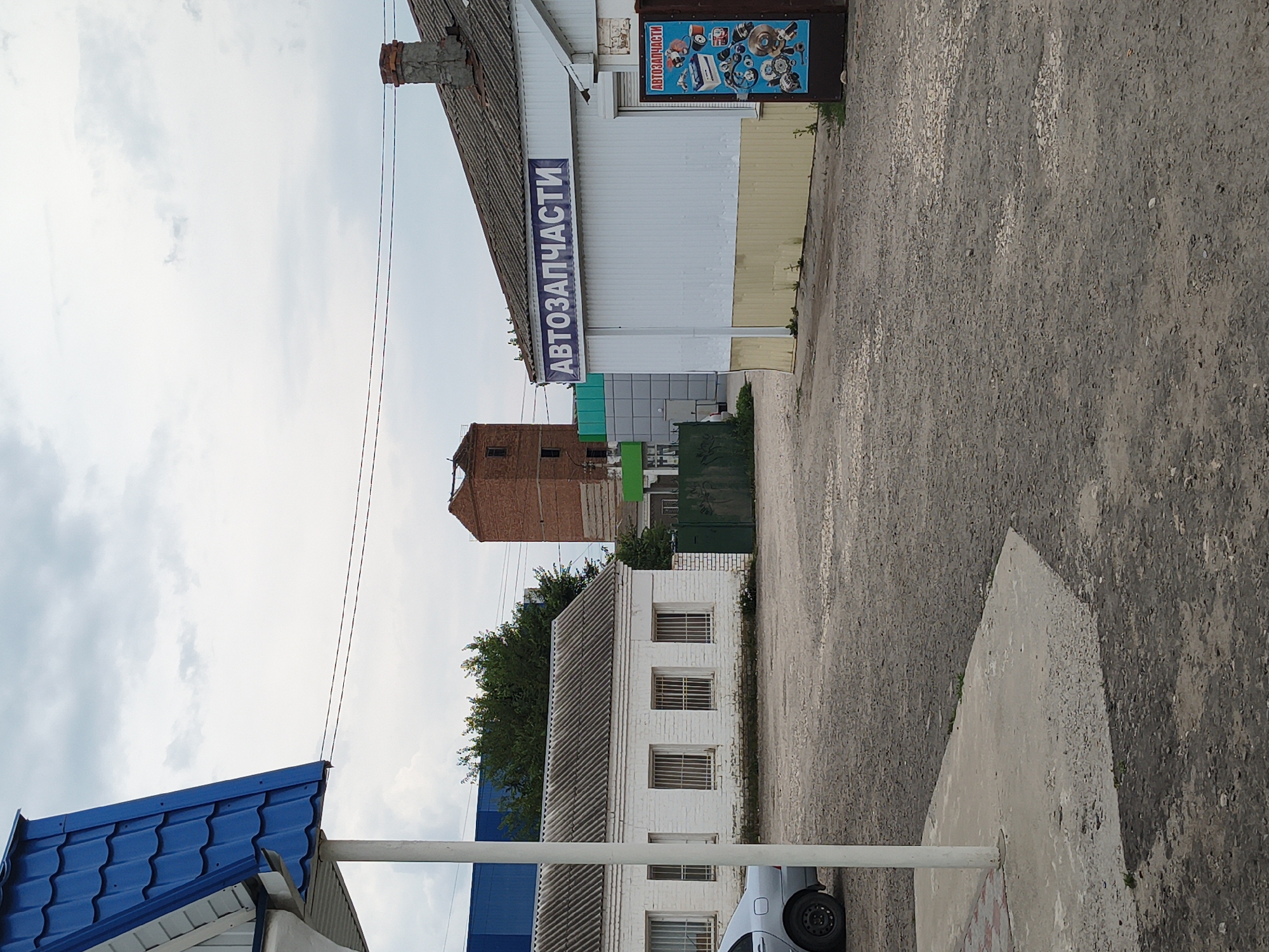The image depicts an outdoor scene in Mikhaylovka, Russia, likely a commercial area. In the foreground, a gravel lot surrounds a concrete pathway and a white pole. To the left, a white building with barred windows is visible, partially obscured by a blue-roofed structure. Beyond this, a silver car is parked. In the mid-ground, a long, white, corrugated building with a sign reading "АВТОЗАПЧАСТИ" (Auto Parts) is prominently displayed. To the right of this building, a vertical sign with the same text and an assortment of car parts is mounted. Further in the background, a tall, reddish-brown brick structure with exposed rebar stands under a cloudy sky, and other industrial or commercial buildings are present. Green fencing and trees are also scattered throughout the scene. The overall impression is one of a somewhat utilitarian commercial district.