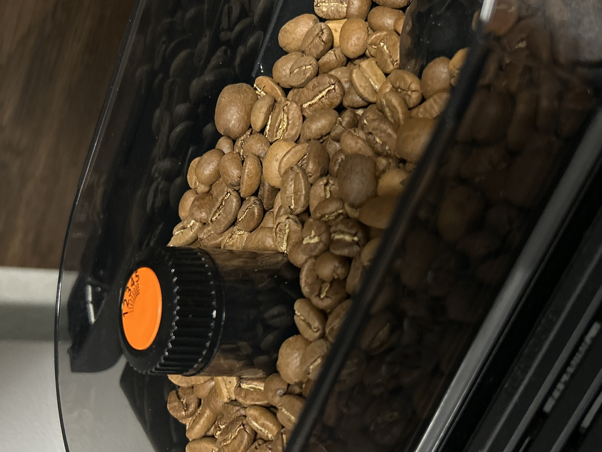 The image shows a close-up view inside a coffee grinder's hopper, filled with roasted coffee beans. The hopper appears to be made of dark plastic. The beans are light brown and show the characteristic split. A black adjustment knob with an orange label is visible, indicating the grind setting. There is no indication this is in St. Petersburg, Russia.