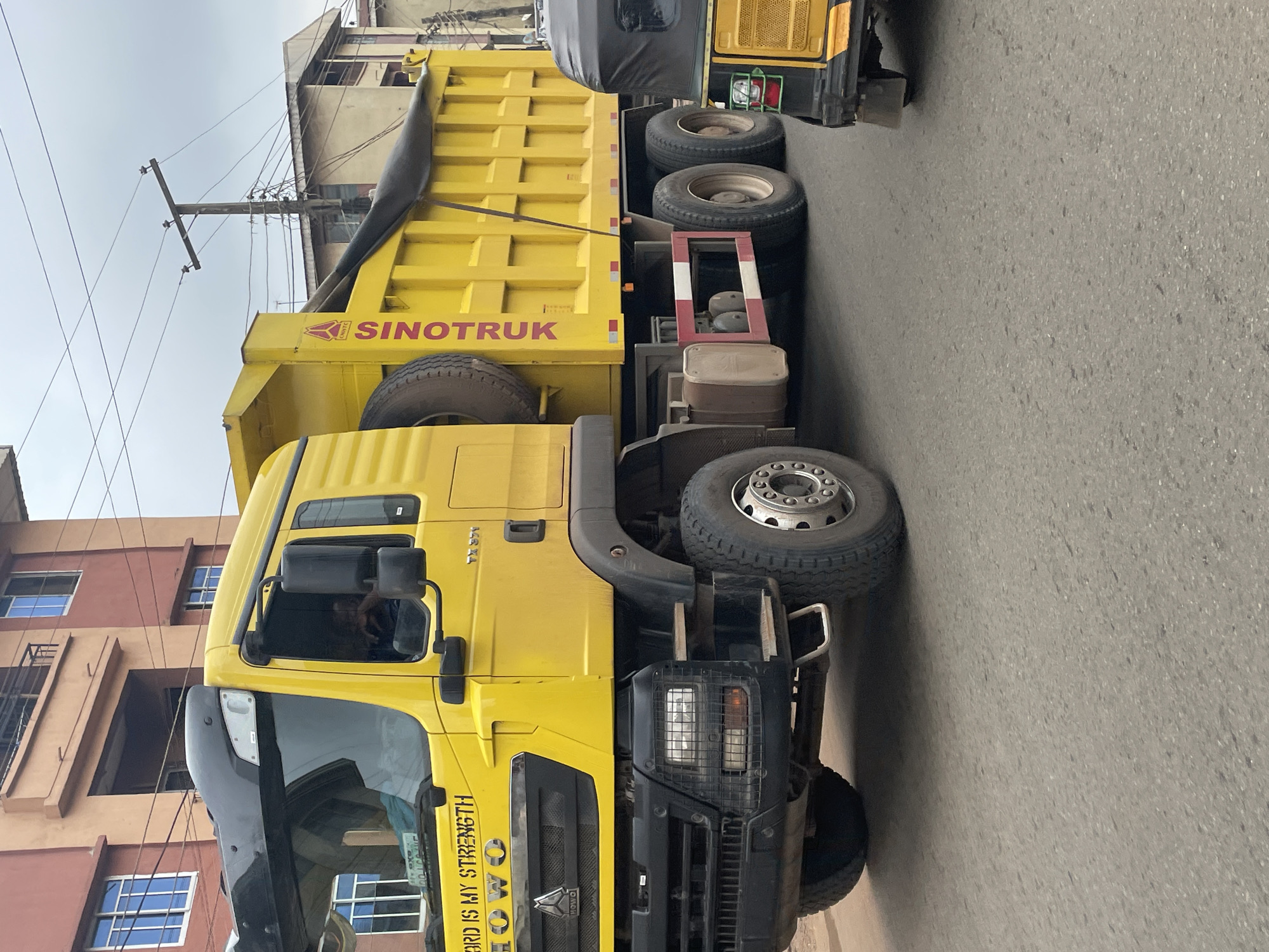 This is a medium-shot, eye-level, outdoor photograph taken on a street in Nkpor, Nigeria, on an overcast day. The primary subject is a large, bright yellow Sinotruk dump truck parked on the right side of the street. The truck features a black cab with a large windshield, and the side of the dump body is emblazoned with the Sinotruk logo and text. There is a spare tire mounted on the side of the dump body. The truck is seen from the front quarter, with its front and rear wheels visible.
To the right of the dump truck, partially visible, is a three-wheeled vehicle, possibly an auto-rickshaw, also yellow and black. It appears to be stationary.
The background shows a multi-story building with orange and brown facades, featuring multiple windows. Overhead power lines and a utility pole are visible against the grey sky. The street itself is asphalt with visible tire marks. The scene appears to be a typical urban street environment in Nigeria, with ongoing vehicular traffic.