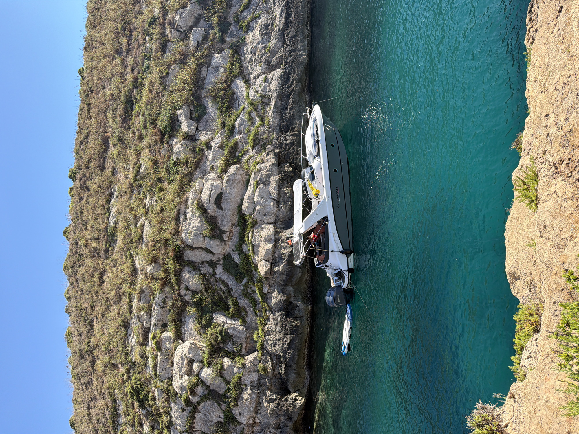 This media file captures a scenic view of a boat docked near a rocky cliff face in Xlendi, Malta. The boat, a sleek white and dark grey vessel, appears to be a motor yacht. A paddleboard is tied to the back of the boat, suggesting recreational activity. Two individuals are visible on the deck of the boat, enjoying the day.
The scene is set against a backdrop of rugged, vegetated cliffs and crystal-clear turquoise water. The sun is shining brightly, casting dappled light on the water's surface and illuminating the textures of the rock. The clear blue sky indicates fair weather, likely during the day. The water's transparency allows for a view of the seabed in some areas. The image is oriented sideways, with the sky on the left and the sea on the right, presenting the scene from an elevated, possibly drone, perspective. The overall atmosphere is one of tranquility and leisure, typical of a Mediterranean coastal location. The name "CALIONBOATS" is visible on the side of the boat.