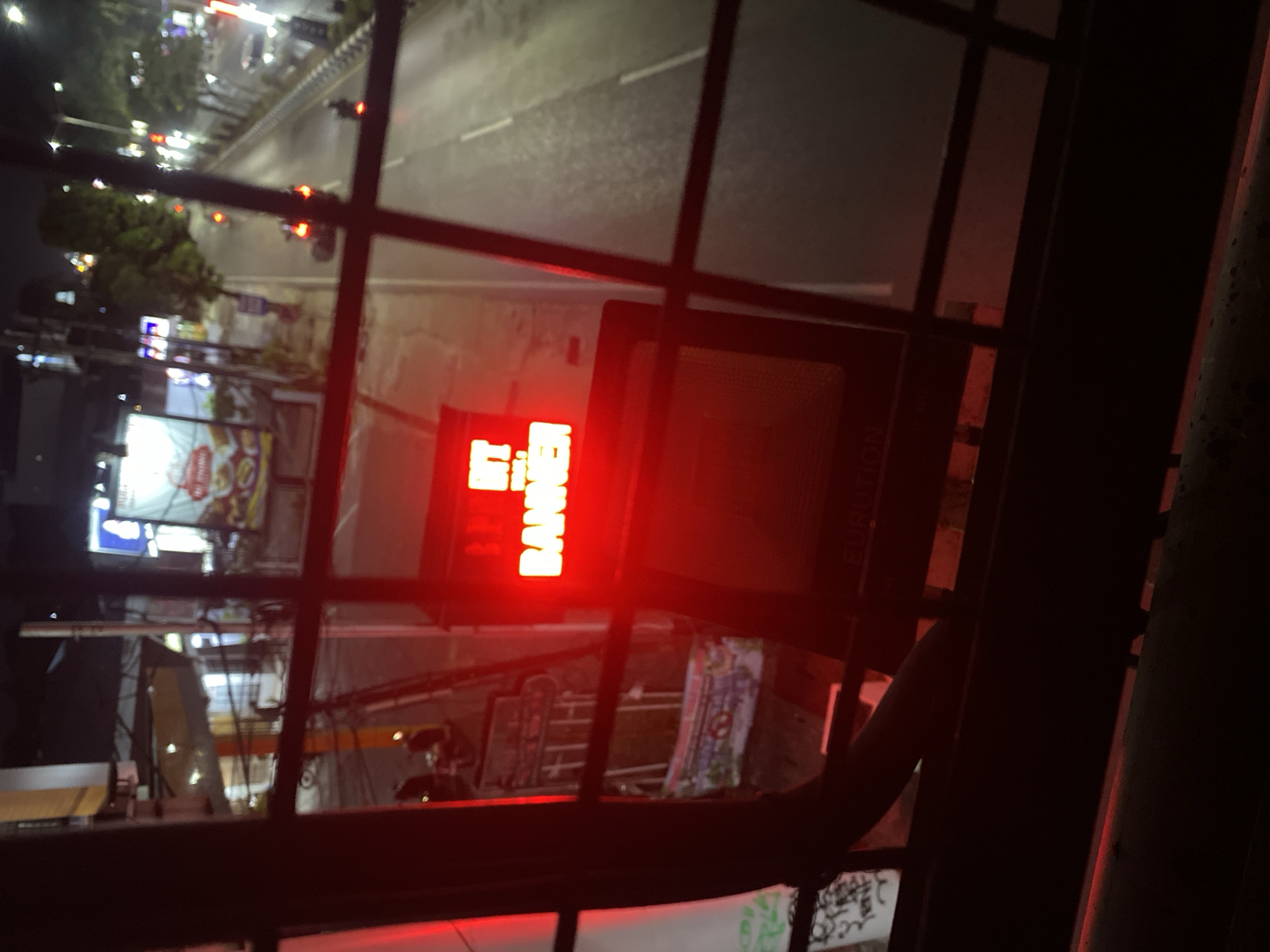 An elevated, night-time shot captures a street scene in Banjarbaru, Indonesia, viewed through a metal grid. Dominating the foreground is a red LED sign displaying the words "HIT BANNER" in bright, block letters. Below the sign, a dark object with a textured surface is visible, possibly a speaker or electronic device.
The background reveals a busy street at night, with vehicles, including motorcycles and cars, illuminated by streetlights and their headlights. Trees line the side of the road, and various buildings and commercial signs are sparsely visible in the distance. The overall impression is of a typical urban environment at night, with the prominent red sign serving as a central visual element.
