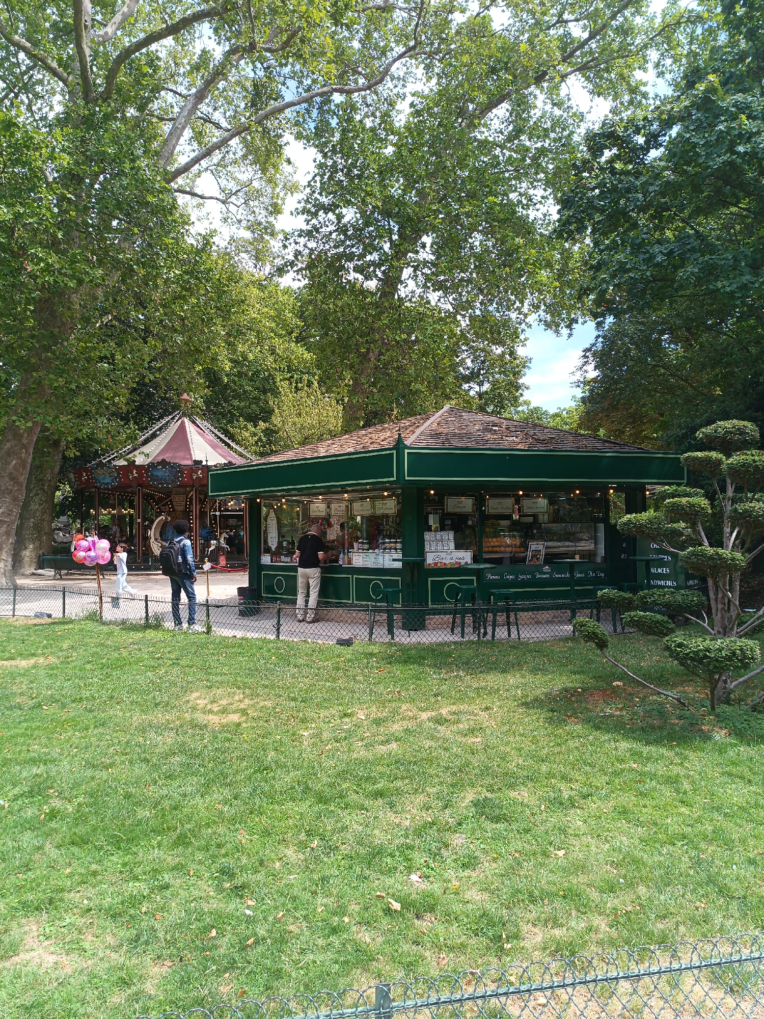 The image depicts a scene in a park, likely in Paris, France, given the presence of a carousel and a concession stand. In the foreground, a lush green lawn is bordered by a wire fence. Behind the fence, a green concession stand with a wooden roof is situated, serving food and drinks. Several people are visible, including a man ordering at the stand and another person waiting. A carousel, a classic amusement ride, is visible in the background, with its colorful roof and decorative elements. Several trees with dense foliage surround the area, providing shade and enhancing the park-like atmosphere. The overall scene suggests a leisurely day in a public park, with families and individuals enjoying recreational activities.