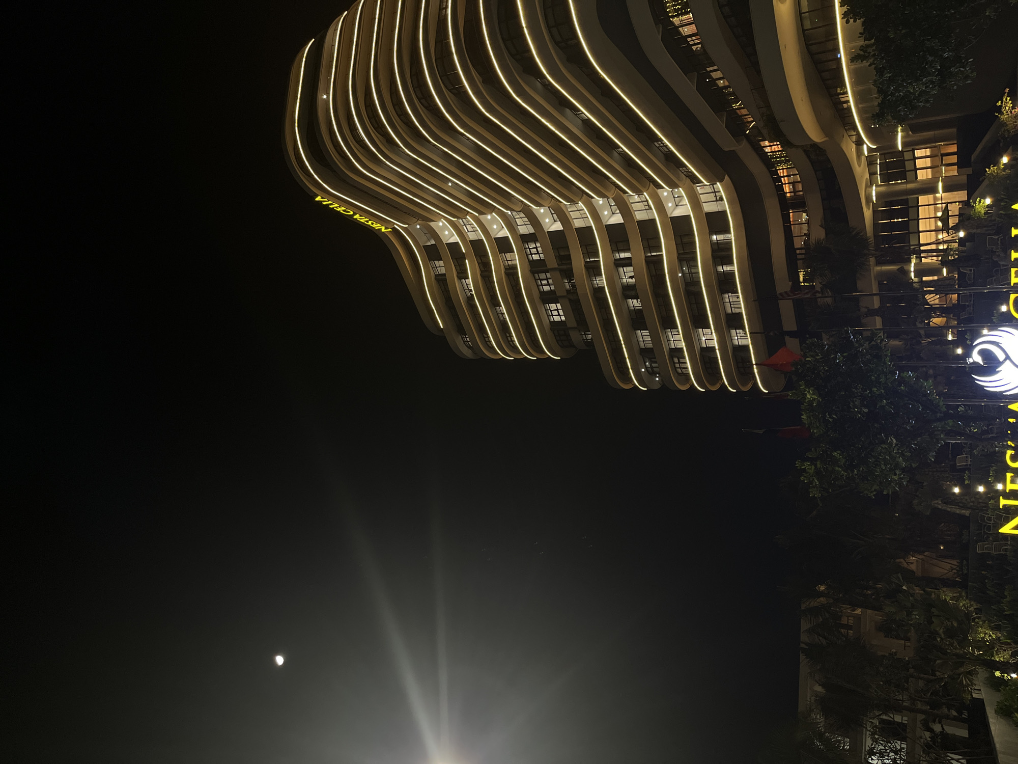 The image depicts a nighttime scene in Đà Nẵng, Vietnam. A multi-story building with a unique, undulating facade is illuminated with linear lights outlining its curved balconies. The building is identified by the signage "NESTA CELIA" in bright yellow letters. Additional signage with a logo is visible on the right side of the building. Below the building, trees and some ambient lighting are visible. In the bottom left corner, a faint moon is visible in the night sky. The rest of the sky is dark.