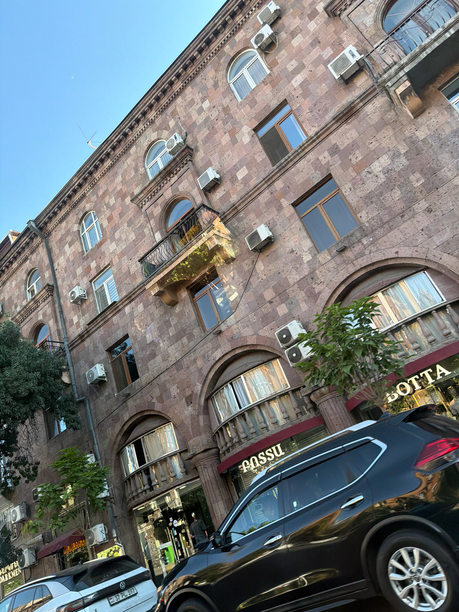 The image shows a street view in Yerevan, Armenia, with a focus on the facade of a stone building. The building has multiple stories, each with numerous windows, some arched and some rectangular, adorned with balconies. Several air conditioning units are mounted on the exterior walls. On the ground floor, shops are visible with signage including "BOTTA" and "FOSSU". A black Nissan SUV and a white Volkswagen SUV are parked on the street. Green trees are present in front of the building. The sky is clear and blue, suggesting daytime.
