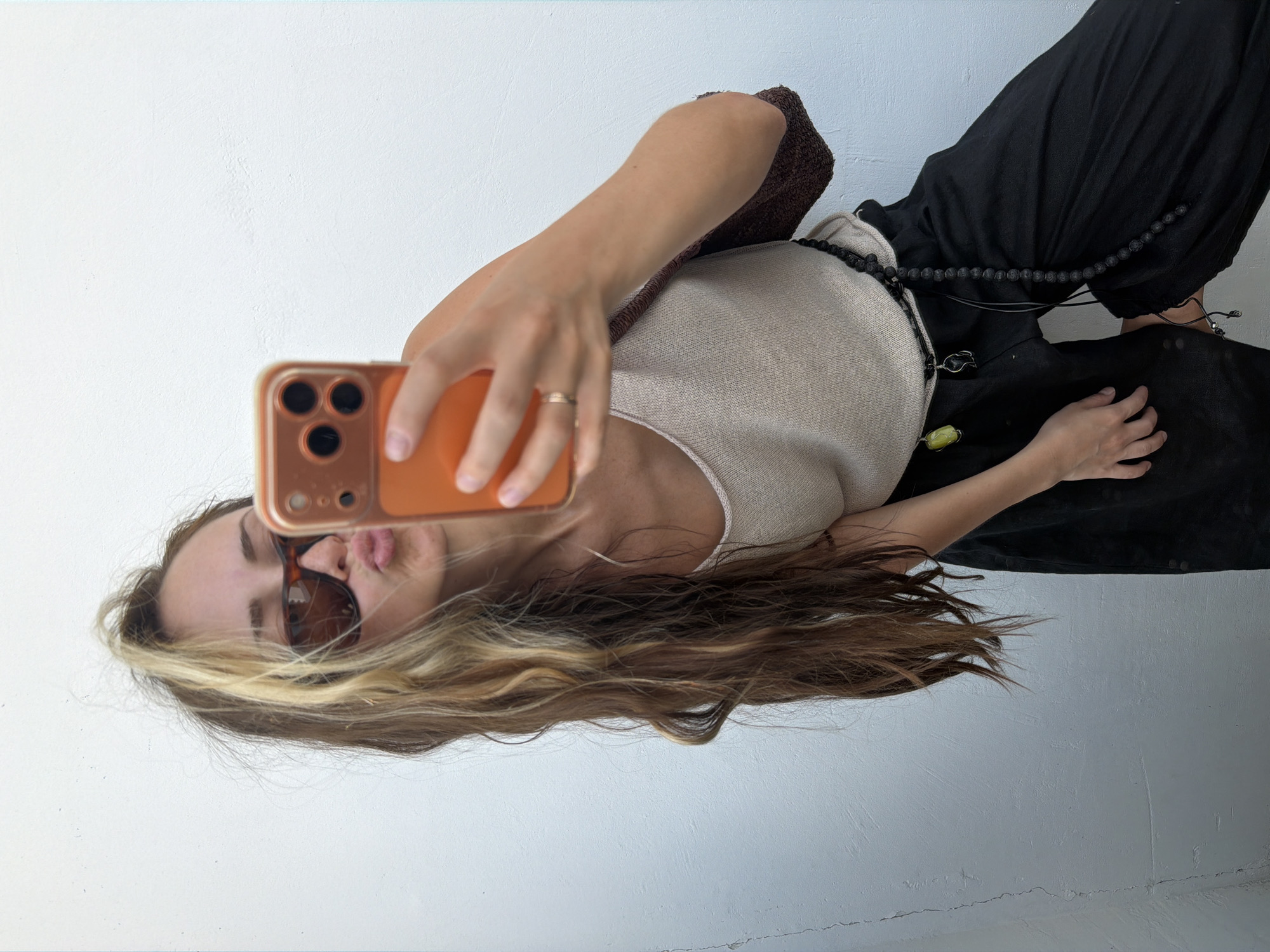 An adult female is taking a selfie with a mobile phone. She is wearing sunglasses and a beige tank top, with black pants and a beaded belt. The background is a white wall. The image does not contain any information about the location in Moscow, Russia.