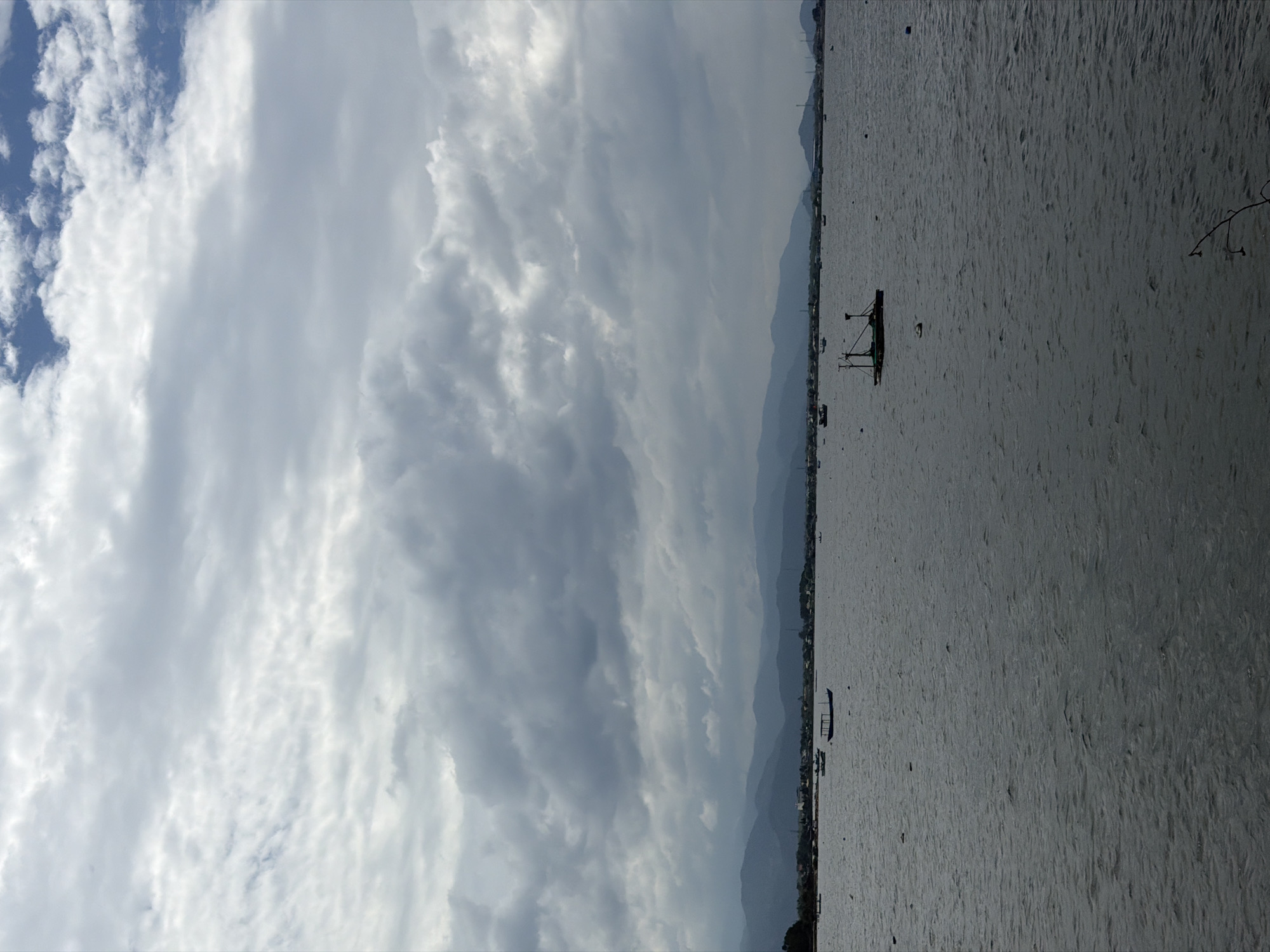The image depicts a serene coastal or bay landscape under a heavily clouded sky. When correctly oriented, the upper portion of the frame is dominated by a vast sky filled with thick, scattered gray and white clouds, with glimpses of clear blue sky visible towards the top left. Below the sky, a series of dark, undulating mountain ranges stretch horizontally across the mid-ground, suggesting a distant landmass. At the base of these mountains, a faint outline of a settlement or village can be discerned along the shoreline.
The lower half of the image is occupied by a wide expanse of water, which appears calm but shows subtle ripples on its surface, reflecting the muted light from the overcast sky. Several boats are visible on the water. A prominent dark boat, featuring an A-frame-like structure, is positioned towards the right-center of the water. Another smaller, longer boat is seen further to the left. More distant, indistinct shapes suggest additional vessels scattered across the water. A bare, dark branch extends into the frame from the far right edge, close to the water.
No people are visible in the scene. The weather appears to be overcast but bright, indicating daytime with soft, diffused light. The overall mood conveyed is one of tranquility and vastness. The presence of traditional boats and mountainous coastlines is consistent with the provided location context of Bac Cam Ranh Ward, Vietnam. There is no visible text in the image.