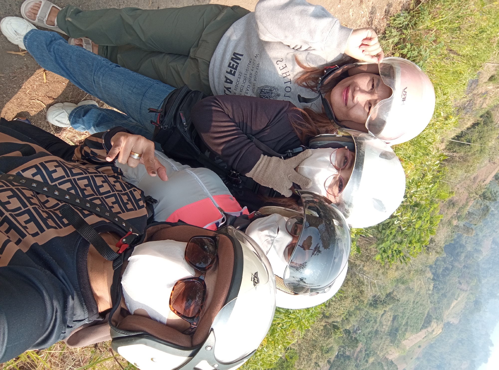 Four individuals, three women and one whose gender is indeterminate, are taking a selfie on a dirt path with a mountainous, vegetated landscape in the background. All individuals are wearing helmets and face masks, and two are wearing sunglasses. The person in the foreground on the right wears a black long-sleeved shirt with a patterned design in brown and black. The person to their left wears a gray jacket with pink accents and a black long-sleeved shirt underneath. The person in the middle wears a black long-sleeved shirt and blue jeans. The person on the far left wears a light gray sweatshirt with text and graphics and green pants. The location is identified as Pandansari Lor, Indonesia, which is a rural area with hilly terrain and vegetation. The image captures a casual moment during an outdoor activity, likely a motorcycle ride given the helmets.