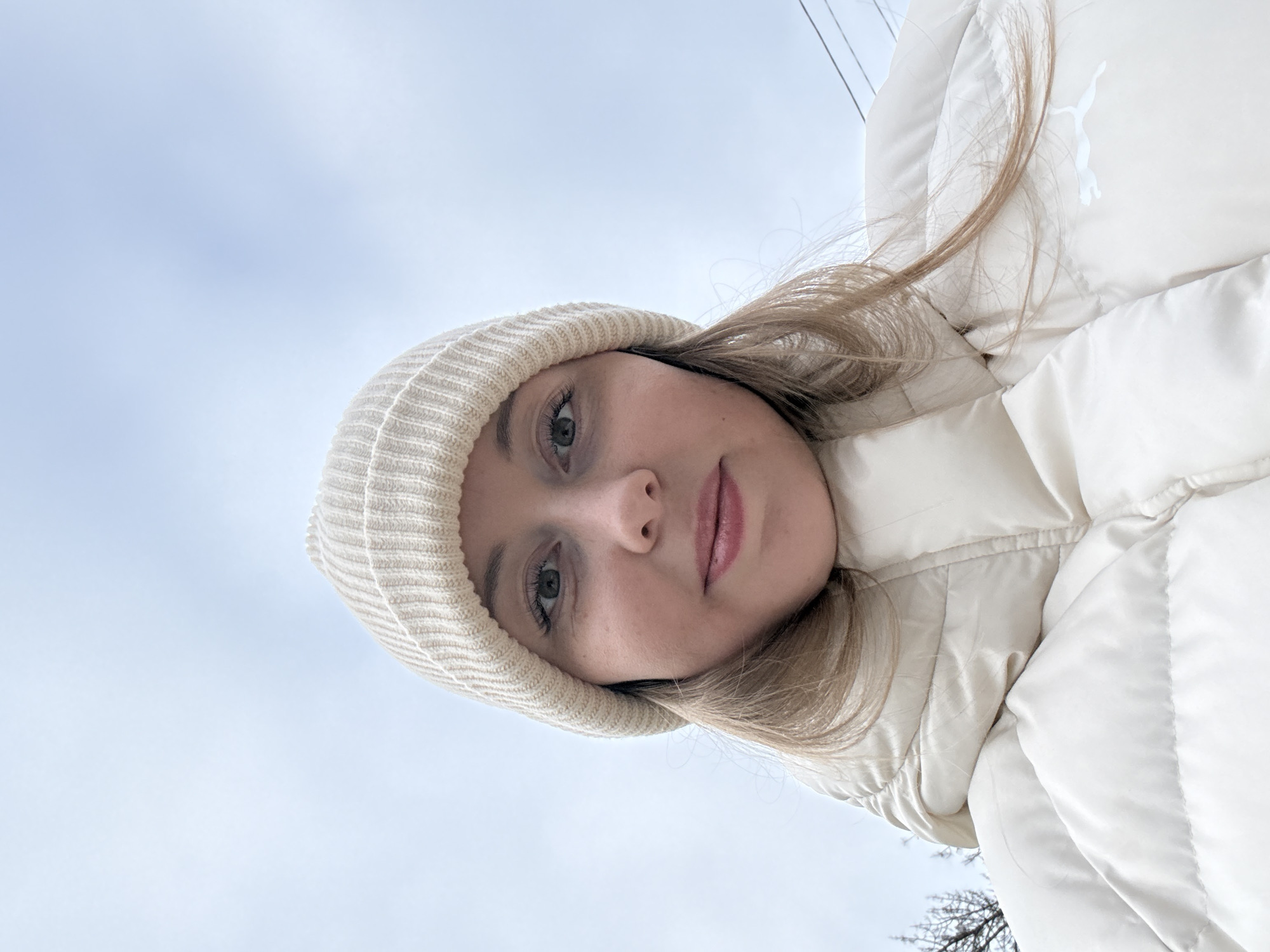 A young woman wearing a cream-colored knit hat and a matching puffer jacket is looking directly at the camera. The background consists of a light blue and cloudy sky, with some faint power lines visible. A portion of bare tree branches is visible in the lower right corner. The woman's jacket has a white Puma logo on the upper right side. The location is in the city of Unknown, Unknown.