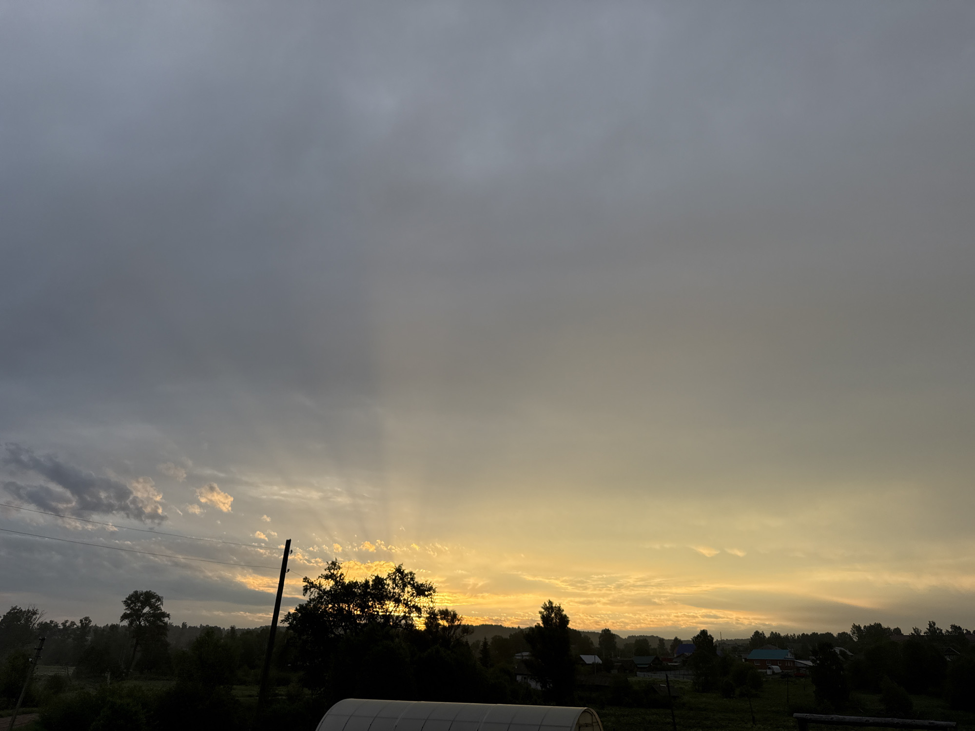 The image depicts a landscape at dusk or dawn in Уральское, Russia. The sky is primarily overcast with a mix of dark gray clouds and areas of orange and yellow where the sun is breaking through. Sun rays are visible through the clouds.
In the foreground, there's a silhouetted treeline and some buildings or structures, including what appears to be a greenhouse. Other buildings, likely houses, are visible in the distance. A utility pole is visible to the left of the center. The overall impression is one of a rural or semi-rural area on an overcast day with the sun either rising or setting.