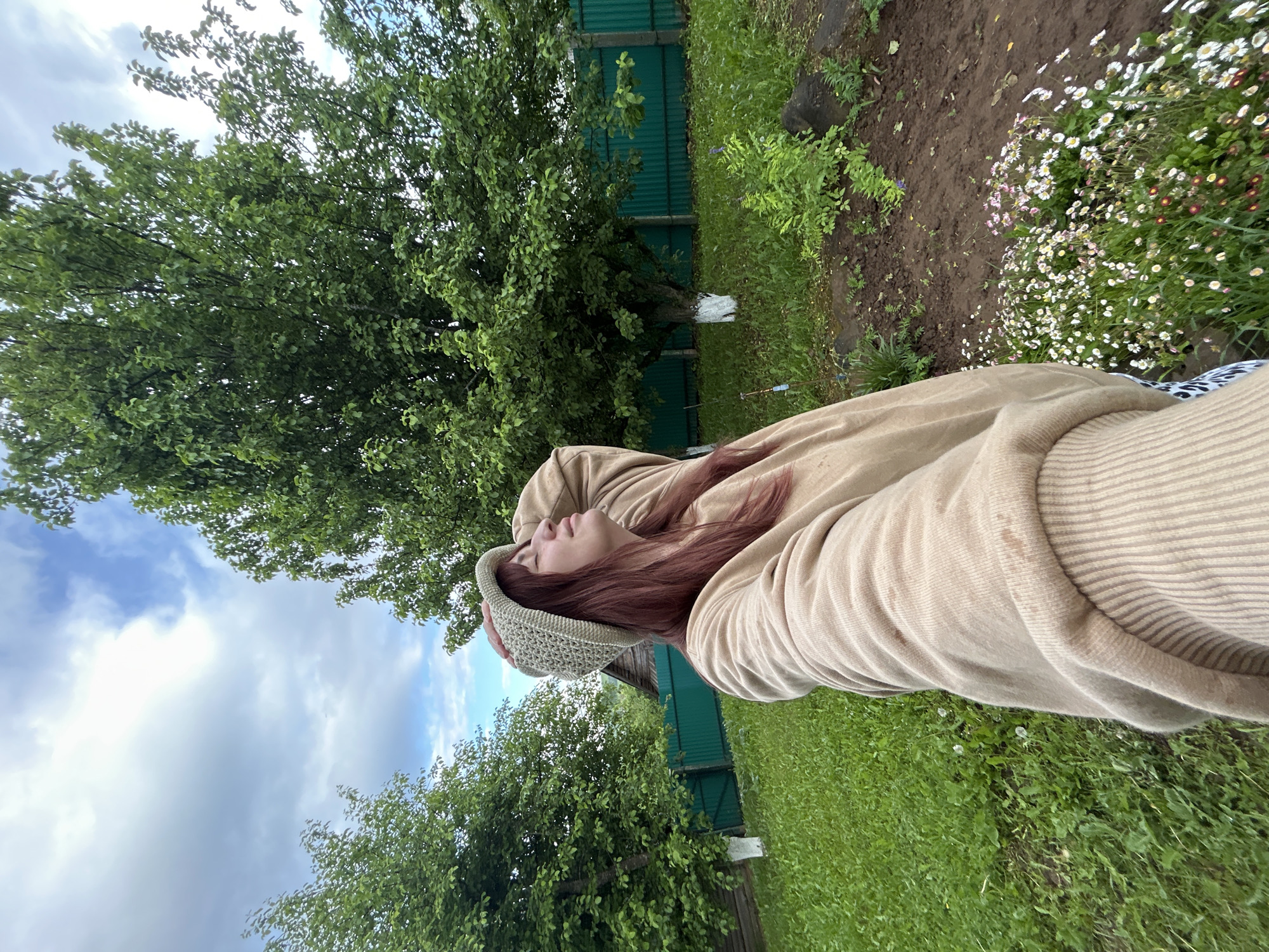The image, captured in a sideways orientation, depicts a person relaxing outdoors in a garden or rural backyard setting, likely in Fokas, Russia. The subject, a person with long reddish-brown hair, is lying down with eyes closed, suggesting a moment of rest or contemplation. They are wearing a light beige or tan hooded sweatshirt and a light green knitted beanie.
The background is rich with natural elements. A large deciduous tree with dense green foliage occupies the left side of the frame, its branches extending towards the top and bottom. The base of the tree's trunk is marked with a distinctive patch of white paint. Behind the tree, a dark green corrugated metal fence is visible. The ground cover varies, with vibrant green grass on the right side and a patch of dark, tilled earth on the left. A cluster of small white and pink daisy-like flowers blooms near the edge of the dirt patch. The sky, seen through the tree canopy, is partially cloudy blue, indicating daytime. No visible text is present. The scene conveys a sense of peaceful natural surroundings.