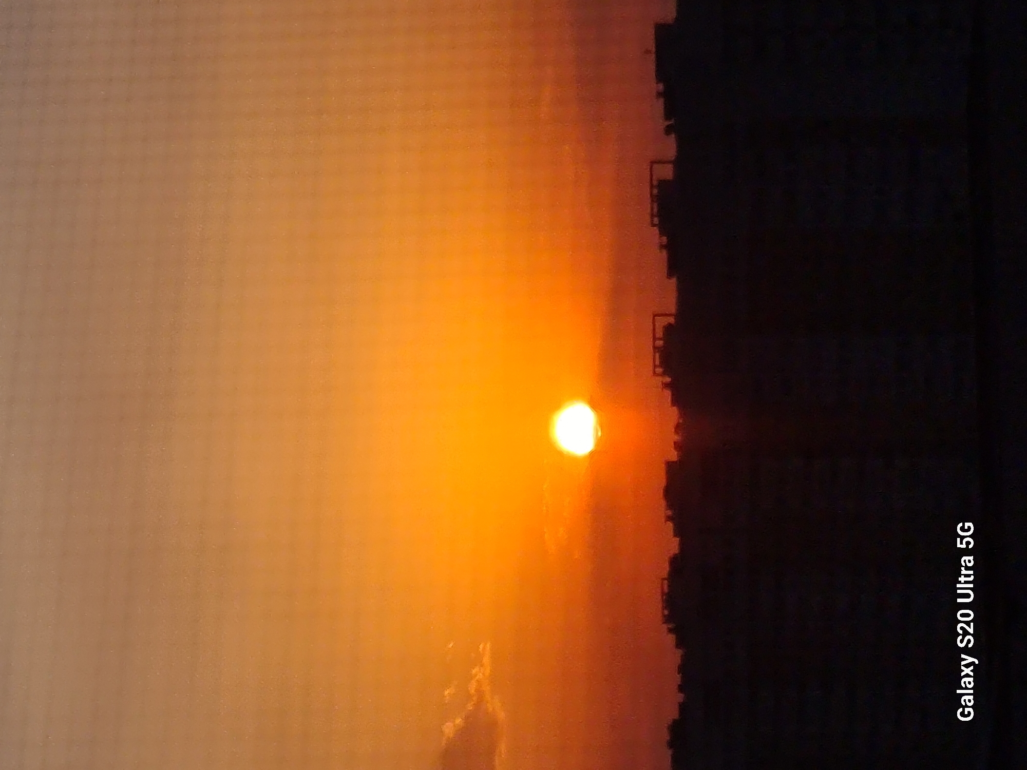 The image shows a sunset over a city, with the sun partially obscured by a thin layer of clouds and appearing as a bright orange orb near the horizon. The sky transitions from a pale orange at the top to a deeper, richer orange closer to the horizon. Silhouetted buildings form a dark, jagged line across the lower portion of the frame. The overall mood is serene and atmospheric, indicative of a city at dusk. This scene is likely from Almaty, Kazakhstan.