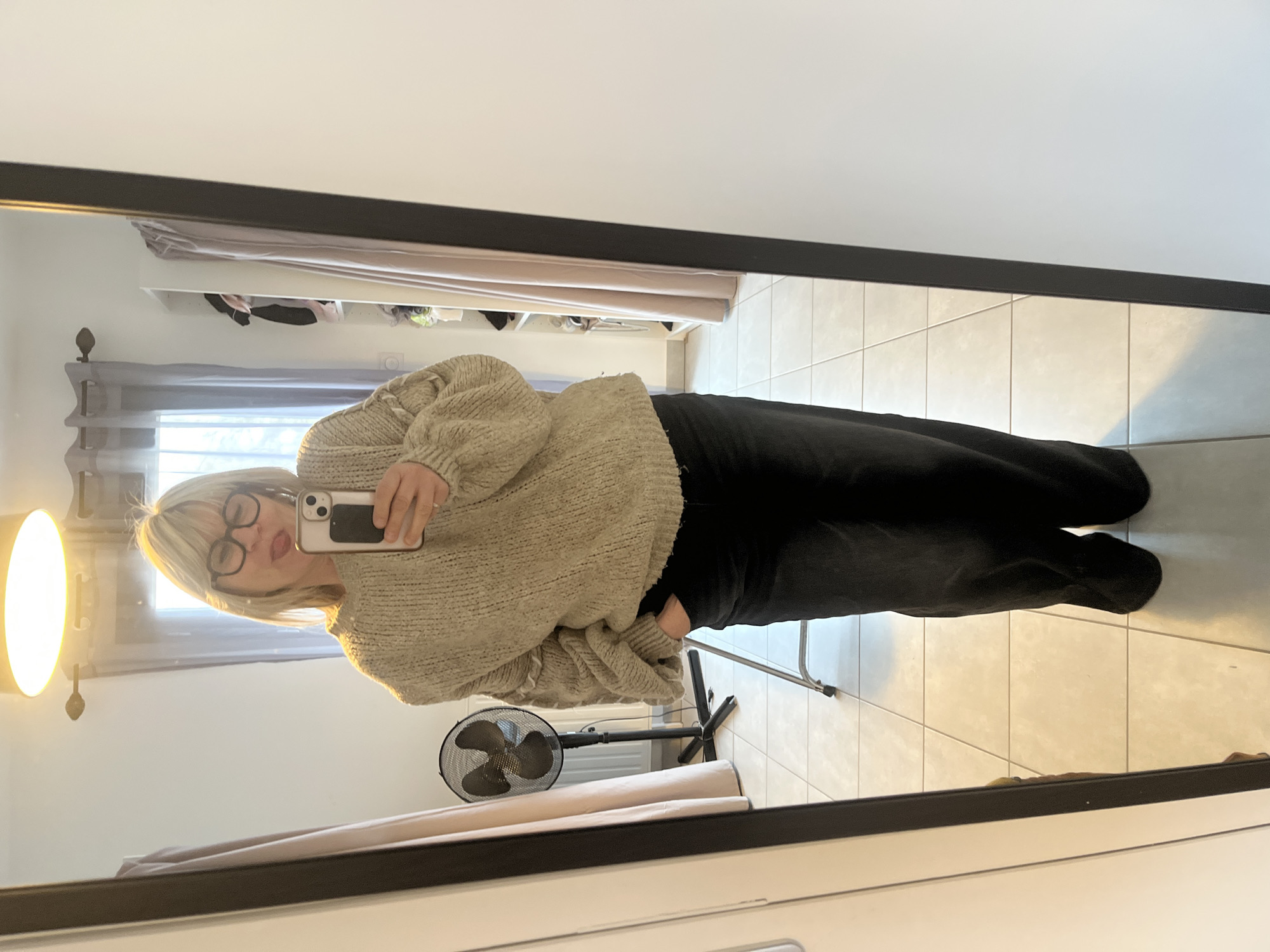 The image depicts an adult female reflected in a full-length, dark-framed mirror, located within an indoor residential setting in Chambœuf, France. The subject, possessing blonde/light-colored hair with bangs and wearing black-rimmed glasses, is dressed in an oversized light beige knit sweater and dark, loose-fitting trousers. She holds a light-colored smartphone with a black grip/accessory in her right hand, extended to capture the mirror selfie. Her left hand is partially inserted into her left trouser pocket. Her tongue is extended. The floor consists of light-colored square tiles. The background, visible in the mirror's reflection, includes a circular ceiling light fixture illuminating the space. To the left, a window with grey curtains, a black pedestal fan, and a white radiator are present. A light-colored metal stand is also visible near the fan. To the right, an open white storage unit displaying various clothing items and additional light-colored curtains are observed.