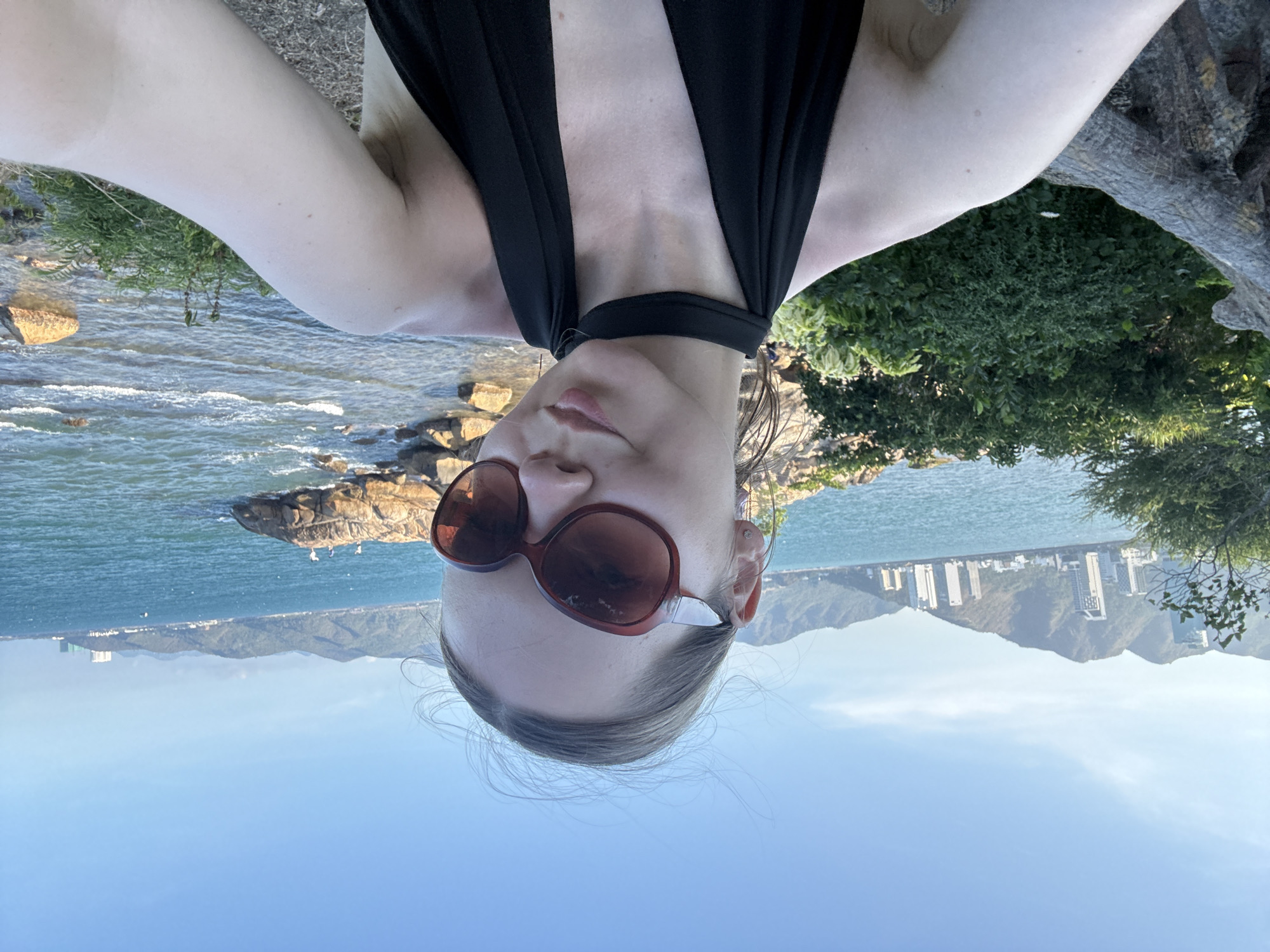 A woman in sunglasses and a black top is taking a selfie. Behind her is a body of water with rocks and vegetation along the shoreline. In the distance are mountains and buildings. It is likely Bac Nha Trang Ward, Vietnam.
