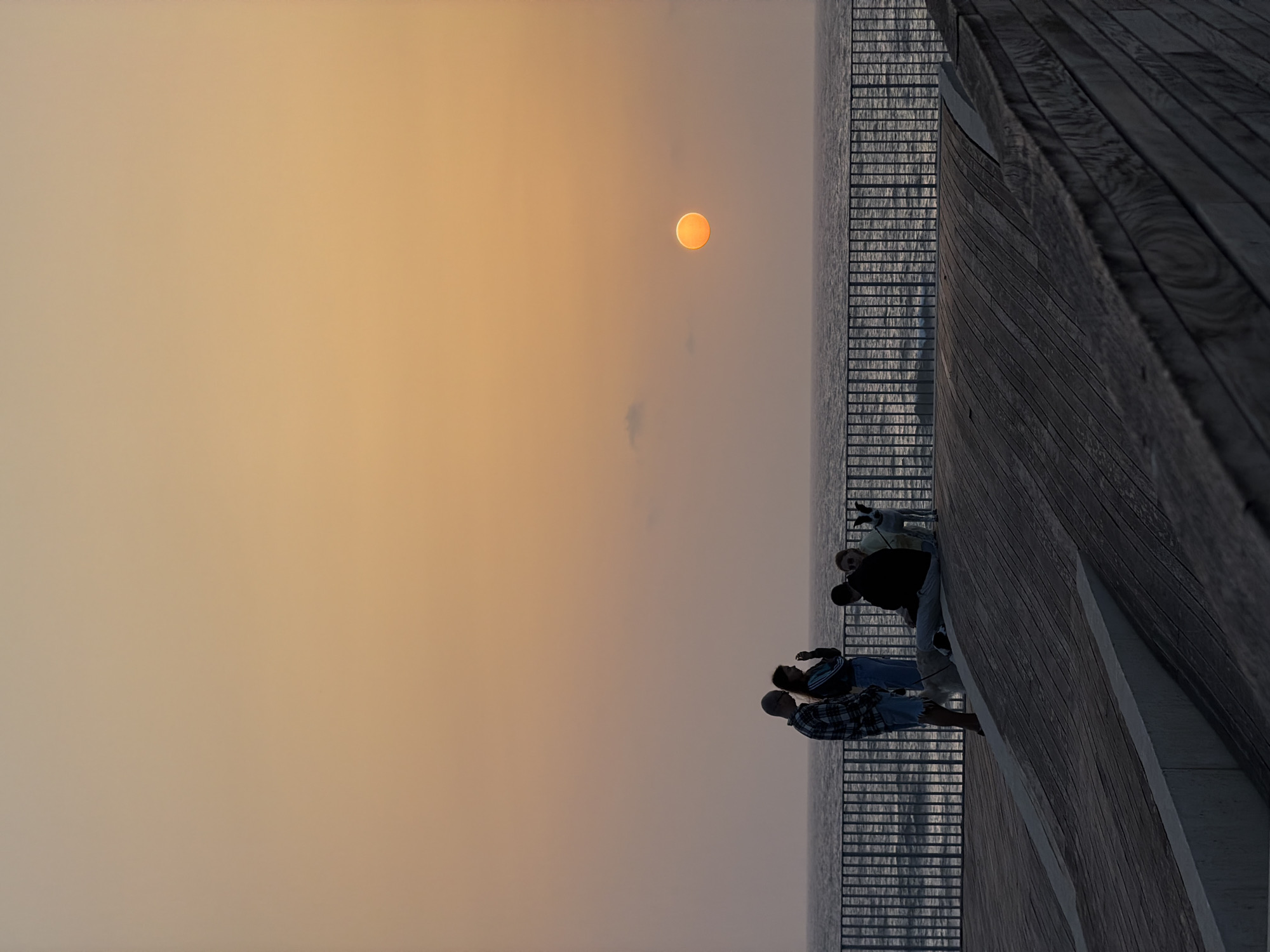 The image displays a tranquil sunset scene over a body of water, likely the Mediterranean Sea, from a modern wooden promenade or pier in Tel Aviv, Israel. The sky, bathed in the soft, warm light of dusk, transitions from a bright orange-yellow around the sun to a muted beige-gray higher up, with a faint, wispy cloud visible near the center.
A prominent, circular orange sun is positioned in the upper left quadrant of the frame, casting a gentle glow. Below the sun, the calm water extends horizontally across the image, separated from the viewer's immediate foreground by a dark, horizontal railing. The water's surface reflects the subdued light, appearing dark and subtly textured.
On the right side of the image, a robust wooden structure, possibly part of a boardwalk or viewing platform, extends into the foreground. Its dark, rich texture and grain are clearly visible. Several people are gathered on this structure, largely silhouetted against the brighter sky and water. They appear to be observing the sunset. Among them, one individual in the foreground wears a plaid shirt and dark shorts, facing towards the water and the sun. Other figures, less distinct, are also present, their stances suggesting contemplation or enjoyment of the view. No discernible text is visible in the image. The overall mood is peaceful and contemplative, capturing the beauty of the twilight hour.