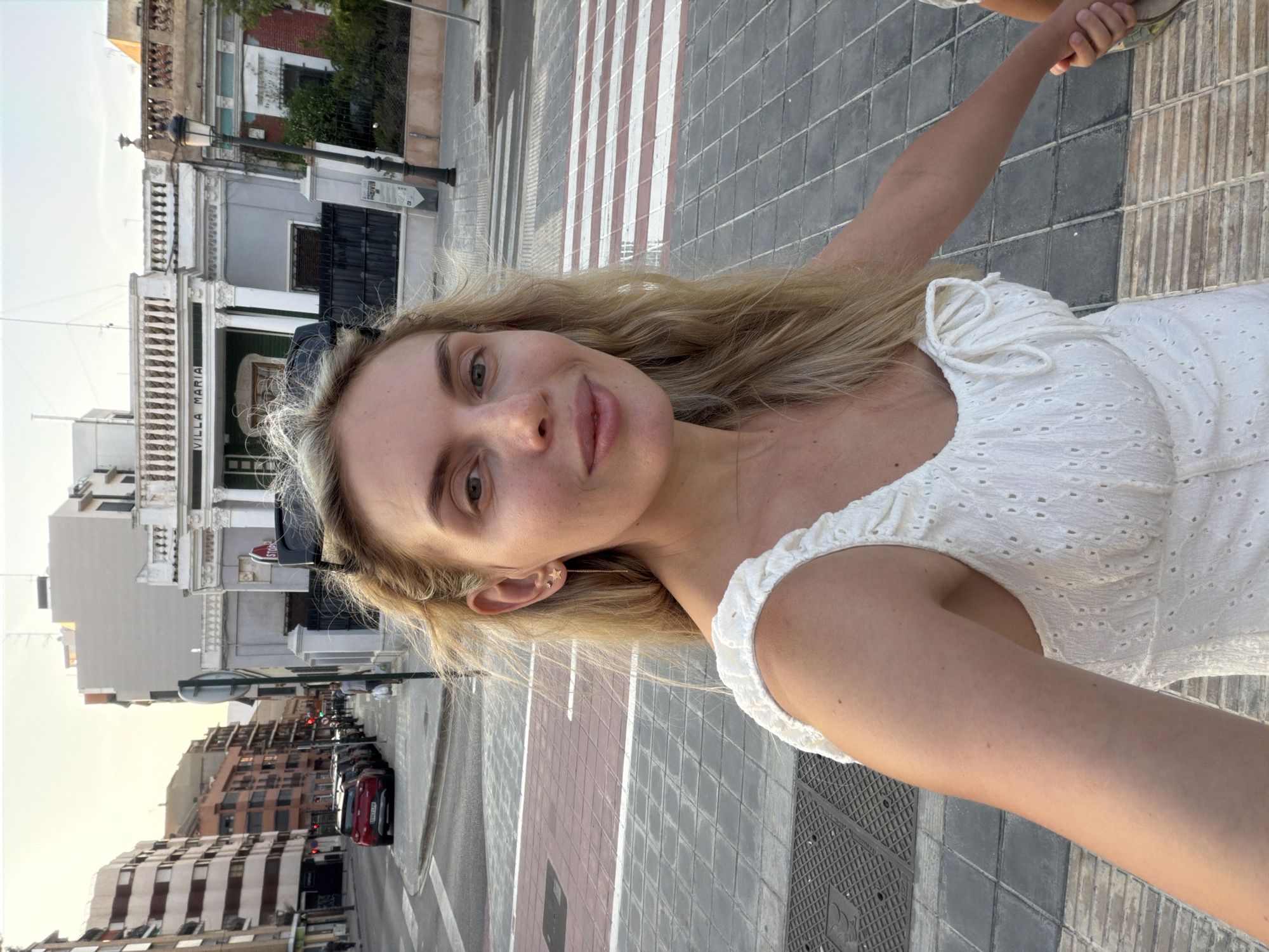 A young woman with blonde hair, wearing a white eyelet dress and star earrings, is taking a selfie on a street in Benimàmet-beniferri, Spain. She is standing on a pedestrian crossing with red and white stripes. Behind her, a street is lined with buildings and parked cars. A building with the name "VILLA MARIA" is visible in the background. The sky is clear, suggesting it is daytime. The woman is holding hands with another person, whose hand is visible on the right side of the frame.
