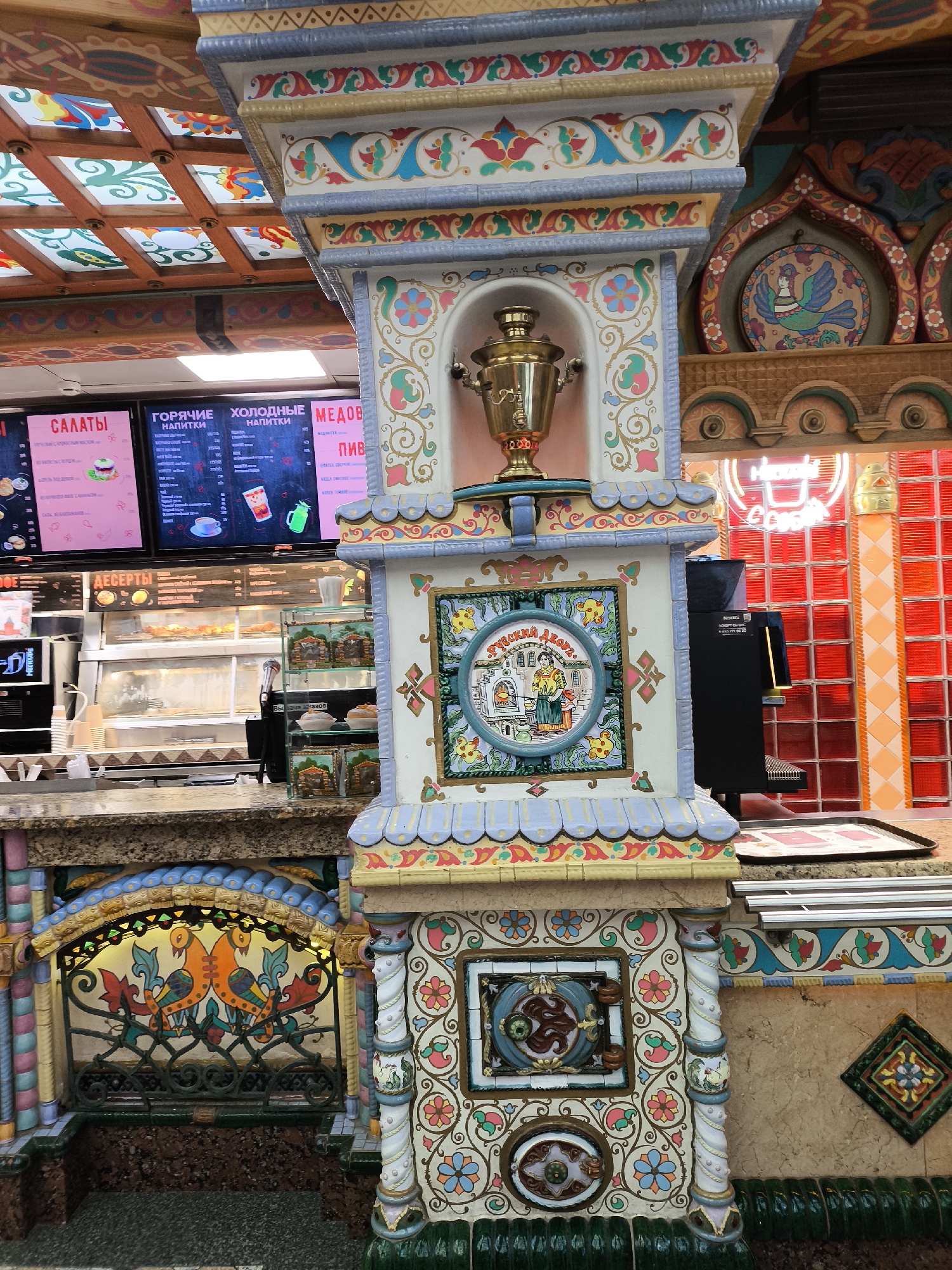 Captured in a vibrant setting of Unknown, Unknown, the image focuses on a decorative food counter adorned in a distinct Russian style. The counter is intricately embellished with colorful tilework and ornate carvings, exhibiting motifs of flowers, birds, and traditional Russian imagery. A golden samovar occupies a niche, adding to the Russian ambiance.
The food counter itself is equipped with digital menu boards showcasing an array of salads, hot and cold drinks, and pastries. Display cases house various food items, indicating an establishment selling quick-service meals. A coffee machine is visible nearby, suggesting coffee is among the beverage offerings.
The background reveals a decorative ceiling with stained-glass-like panels, contributing to the visually rich atmosphere. Neon signs and red block tiles further enhance the setting's lively character. The scene presents a blend of food service with cultural artistry, reflecting the establishment's unique design.