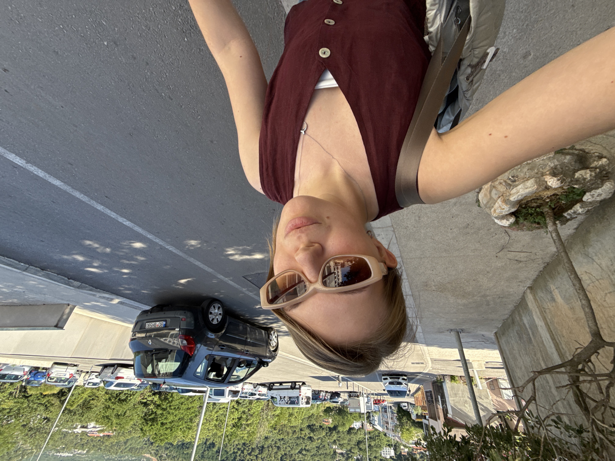 A young woman is captured in a high-angle selfie, standing on a concrete sidewalk adjacent to a street in Budva, Montenegro. She has light brown hair and wears beige rectangular sunglasses, a maroon sleeveless button-down top over a white undershirt, a silver necklace with a small pendant, and a silver hoop earring. A light grey shoulder bag hangs over her right shoulder. To her immediate left, a small tree is planted in a decorative stone pot beside a tall concrete retaining wall. The asphalt road to her right features a white painted line. A dark grey Renault Scenic car, bearing the Montenegrin license plate "BD BH590", is visible on the road, with other parked vehicles including a white Mercedes-Benz and a white van displaying "Zdravlje" text and a graphic. The background shows buildings nestled into a steep, densely tree-covered hillside under a clear sky.