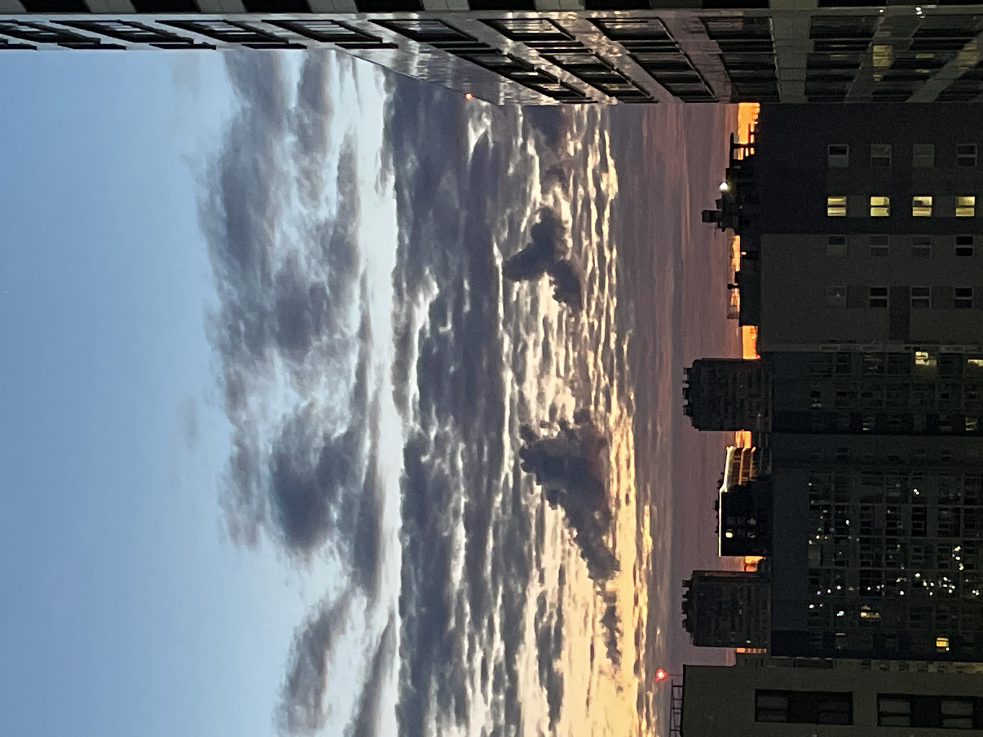This is a vertical, slightly low-angle, outdoor photograph of a city skyline at sunset or sunrise. The foreground is dominated by a vast expanse of clouds, rendered in shades of gray, white, and pale orange. The clouds are textured and layered, suggesting movement and depth.
Flanking the sky are two tall, multi-story buildings. The building on the left is dark with a pattern of regularly spaced rectangular windows. The building on the right is also dark, with numerous illuminated windows casting a warm glow, indicating that people are inside. The architectural style of the buildings suggests an urban environment.
The time of day is clearly indicated by the warm, golden and pink hues in the sky, especially visible at the horizon. The weather appears to be partly cloudy, with the dramatic cloud formations being a prominent feature. There are no people visible in the image. The overall mood is tranquil and slightly dramatic, due to the interplay of light and shadow in the clouds and buildings. The location context is noted as Unknown, Russia.