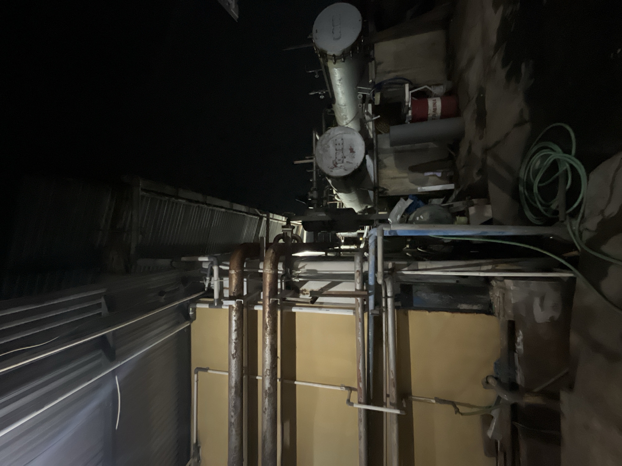 The image depicts an outdoor industrial scene at night, likely in Pekalongan, Indonesia, characterized by the presence of large pipes, tanks, and rudimentary roofing. In the foreground, a coiled green hose lies on the ground. To the left, a yellow-painted wall serves as a backdrop for a network of rusty metal pipes and white PVC pipes, some appearing to be connected to machinery or structures. A corrugated metal roof is visible overhead, supported by beams. To the right, two large, cylindrical tanks are mounted on a raised platform. One tank bears visible markings. A red barrel with the word "PERTAMINA" is positioned near the tanks. The overall environment appears somewhat unkempt, with debris and puddles on the ground. The lighting is dim, suggesting artificial illumination from the facility itself or nearby sources. No people are visible in the frame.