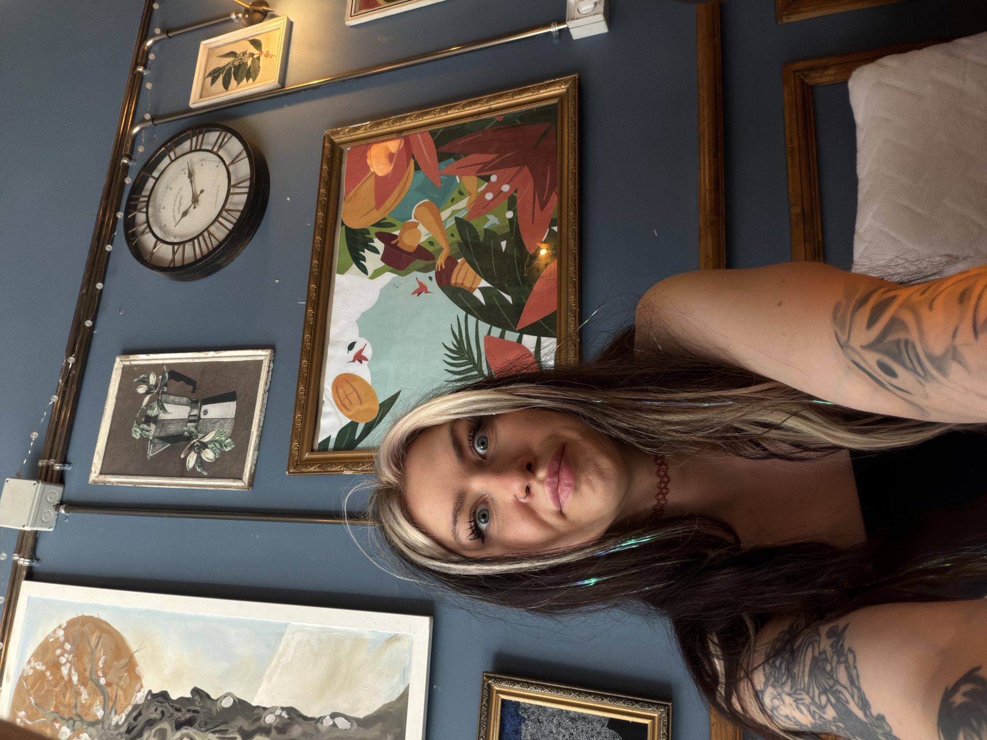 A rotated, tilted shot shows a woman in front of a blue wall decorated with various framed artwork and a clock. The woman has dark hair with a streak of blonde at the front, blue eyes, a nose ring, and is wearing a dark choker necklace. Her arms are visible, both adorned with tattoos. Behind her, on the wall, are several framed pictures and artwork including one in a gold frame with images of flowers and leaves. A large dark clock is on the wall near the pictures. A metallic pipe with hanging lights runs diagonally across the upper left of the frame. The location appears to be a room in Belgrade, Serbia, given the style and decor typical of certain establishments.