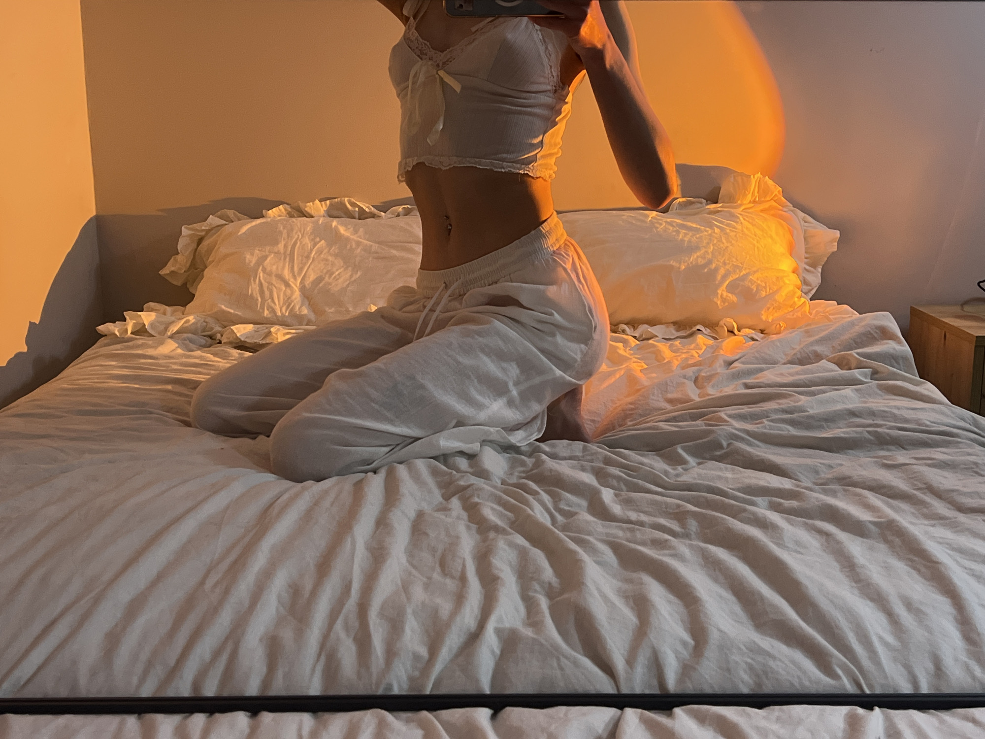 A waist-up indoor shot features a person kneeling on a bed. The person is wearing a white, lacy crop top and white drawstring pants. The location cannot be determined based on the provided image. The bed has rumpled white linens and pillows. A bedside table is visible in the background. The lighting suggests either sunset or artificial lighting designed to mimic the golden hour. The person is holding a phone in their left hand, presumably taking the photograph.