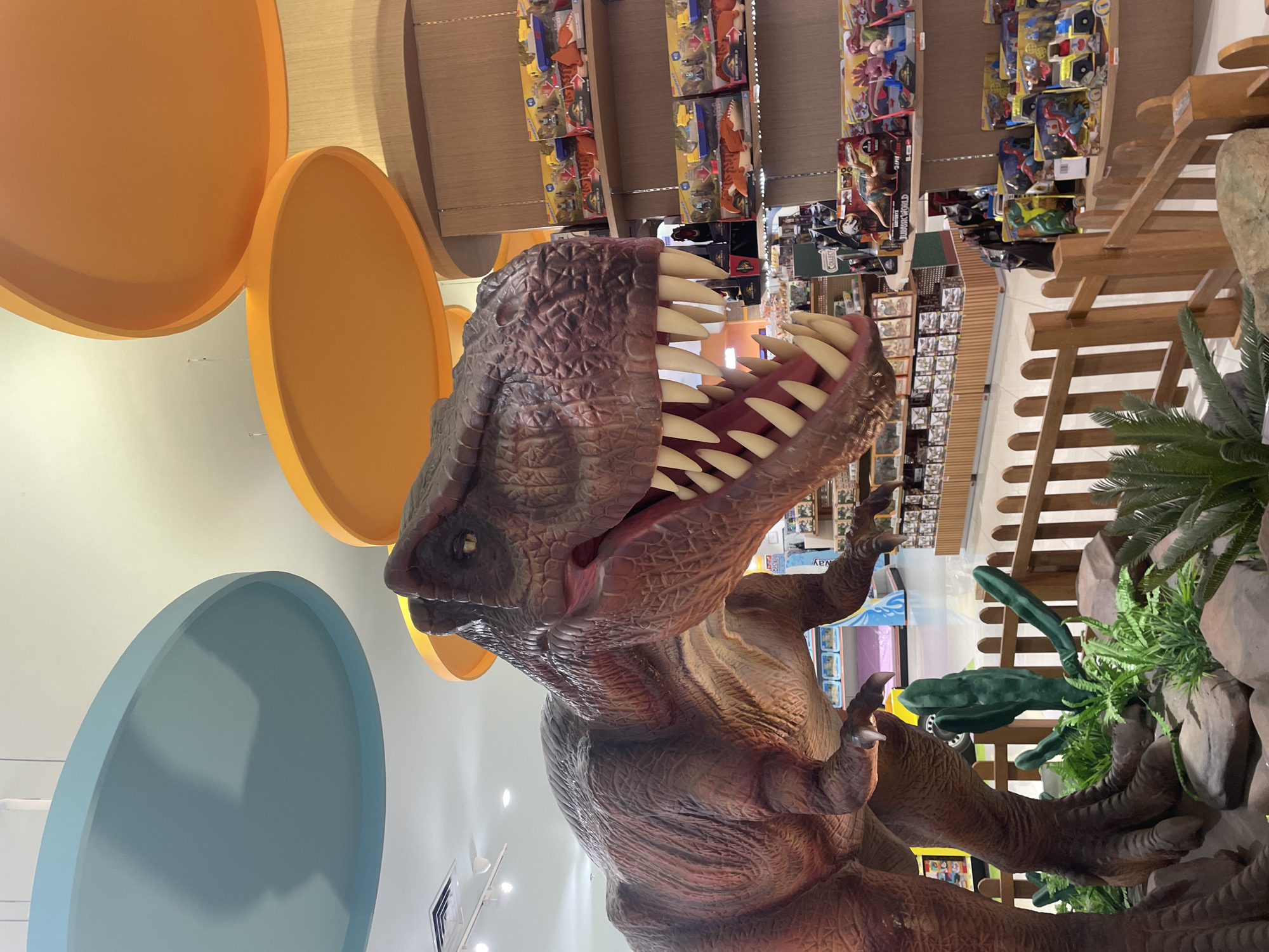 The image shows a life-sized Tyrannosaurus Rex model, likely located in a retail environment. The model is brown and textured to resemble dinosaur skin, with a large open mouth displaying sharp, white teeth. The dinosaur is positioned near displays of toys and other merchandise, suggesting it is a feature in a store. There are also faux rocks and vegetation surrounding the base of the dinosaur. The location is indicated to be Denpasar, Indonesia. There are no people visible in the immediate frame.