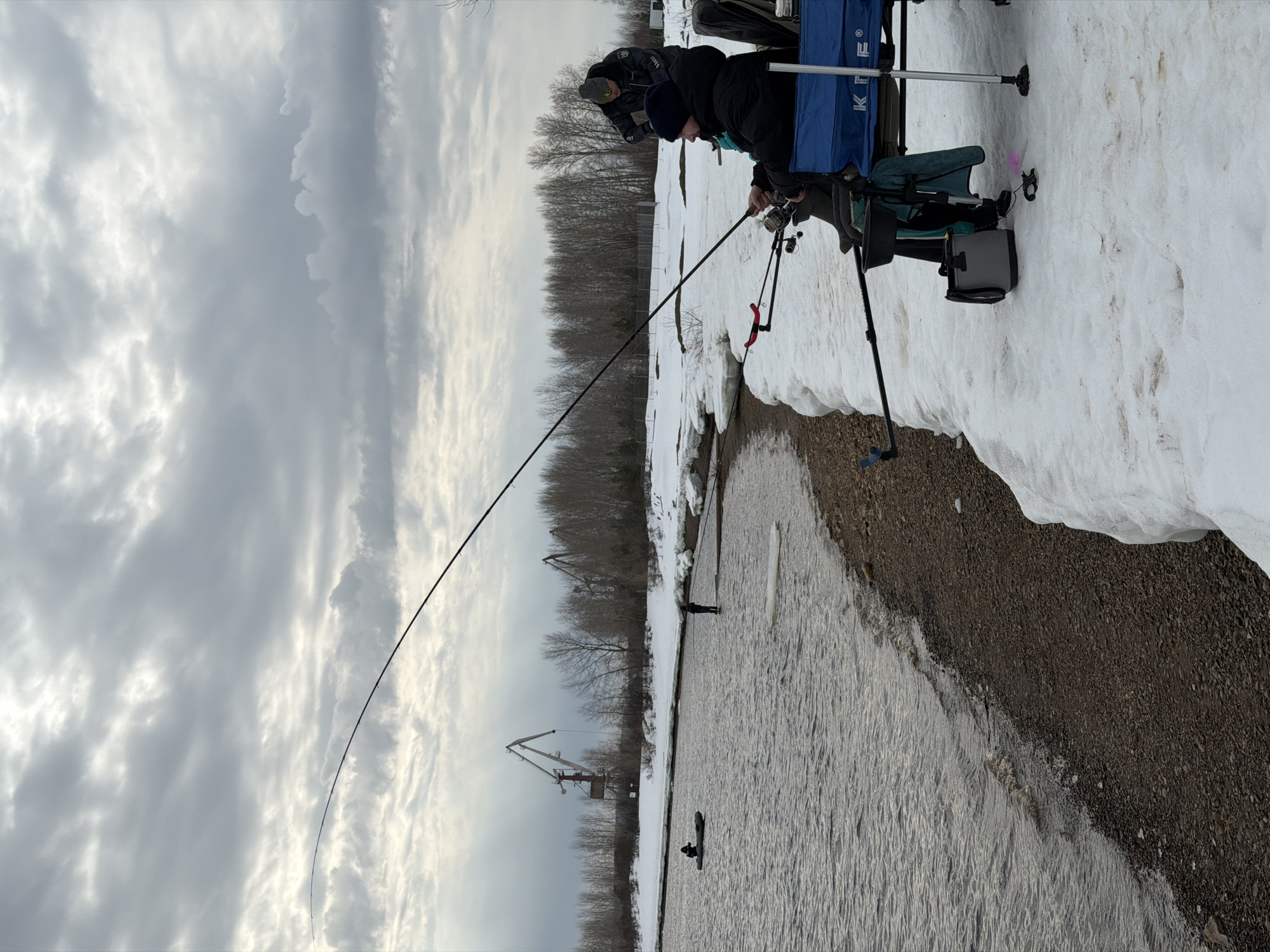The image shows two individuals fishing in Noviy Urengoy, Russia. They are positioned on a snowy bank alongside a body of water. One person is actively fishing, holding a long, curved fishing rod. In the water, there is a person in a kayak and another person standing further away. In the background, there are trees and two large cranes. The sky is overcast with grey clouds.