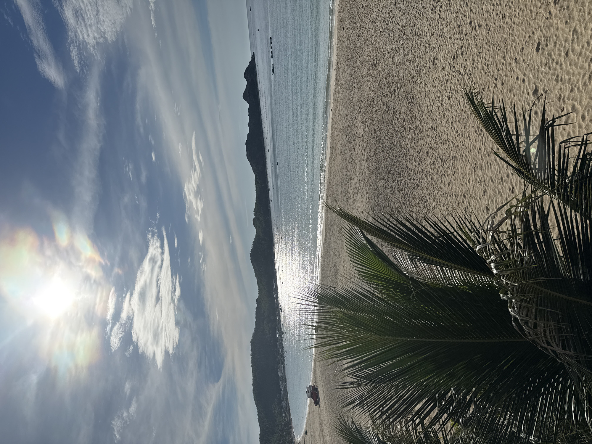A tranquil beach scene unfolds under a bright, partly cloudy sky. The sun, positioned in the upper left, casts a radiant glow with subtle rainbow effects, illuminating the gentle waves of the ocean. A sandy beach stretches across the foreground and midground, leading to the calm turquoise water. In the distance, a cluster of dark, tree-covered hills forms the coastline. A few small boats dot the sea, and a red vehicle with white boxes on its roof is parked on the sand near the water's edge. In the lower left corner, the fronds of a palm tree provide a foreground element, their dark green contrasting with the sun-drenched sand. The overall impression is one of peaceful, tropical serenity.