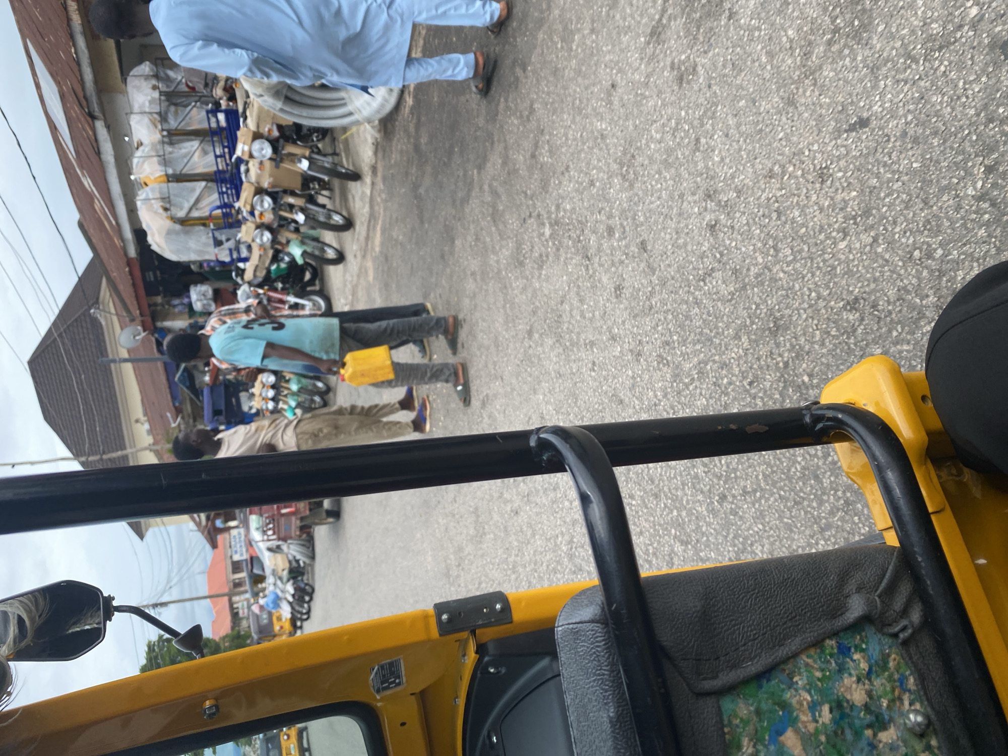 The image depicts a street scene in Jalingo, Nigeria, with a focus on several individuals and a row of motorcycles. In the foreground, a yellow vehicle, likely a three-wheeled auto-rickshaw, occupies the bottom portion of the frame. A person in the distance, wearing a light blue traditional outfit, walks on a paved surface. Another individual, dressed in a light blue shirt and dark pants, is seen carrying a yellow jerrycan. Several motorcycles are parked along the side of the street, some covered with tarps. The setting appears to be a marketplace or a commercial area, indicated by the presence of shops with corrugated metal roofs and the variety of goods displayed. The sky is overcast, suggesting a cloudy day. The overall atmosphere seems to be one of everyday commerce and transportation. No visible text is present in the image.