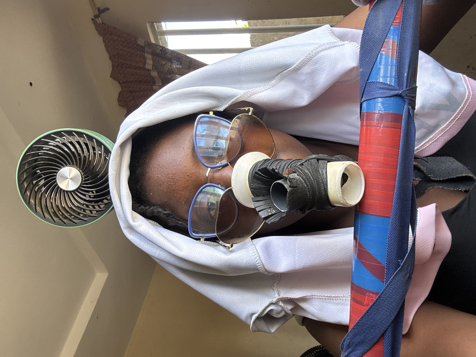 A young Black woman, wearing glasses and a white head covering, is captured in a close-up shot. She appears to be indoors, possibly in Jawu, Nigeria. Above her, a small, green-bladed fan is attached to the ceiling. The woman is holding a cylindrical object wrapped in red, blue, and black tape. A portion of her face is obscured by a white fabric, and she is wearing a mask-like apparatus over her nose and mouth. The background suggests a window with curtains and a wall.
