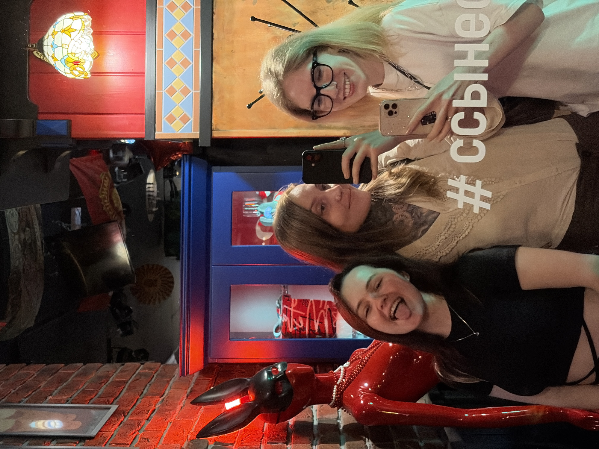 Three young women are posing for a selfie in what appears to be an indoor establishment. The woman on the left is sticking her tongue out, the woman in the middle is taking the selfie with her phone, and the woman on the right is smiling broadly. They are in front of a red, rabbit-like mannequin and a blue cabinet. In the background, there are various decorations including a stained-glass lamp and some hanging banners. The overall scene is dimly lit with red and green lighting. The image contains the hashtag "#ссыне" superimposed on the glass in front of them. There is no information in the image to suggest the location is Izhevsk, Russia.