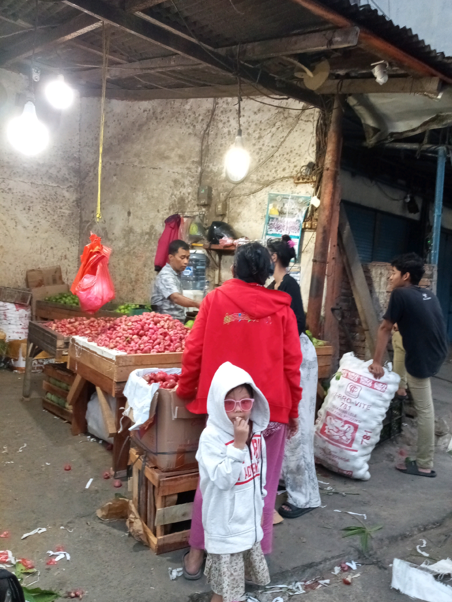 An outdoor market stall in Bekasi, Indonesia, displays a large quantity of pinkish-red fruit, possibly lychees, in wooden crates. A man in a checkered shirt stands behind the fruit, appearing to be a vendor. Several people are present, including a woman in a red hoodie, a young child wearing a white hooded sweatshirt and pink sunglasses, and a young man in a black t-shirt. The market area has a rough concrete floor littered with some debris. Overhead, there are exposed wooden beams and corrugated metal roofing. Electrical wires and light bulbs are visible, along with a hanging red plastic bag. A large white sack with writing is positioned near the young man. The scene suggests a busy, open-air marketplace.