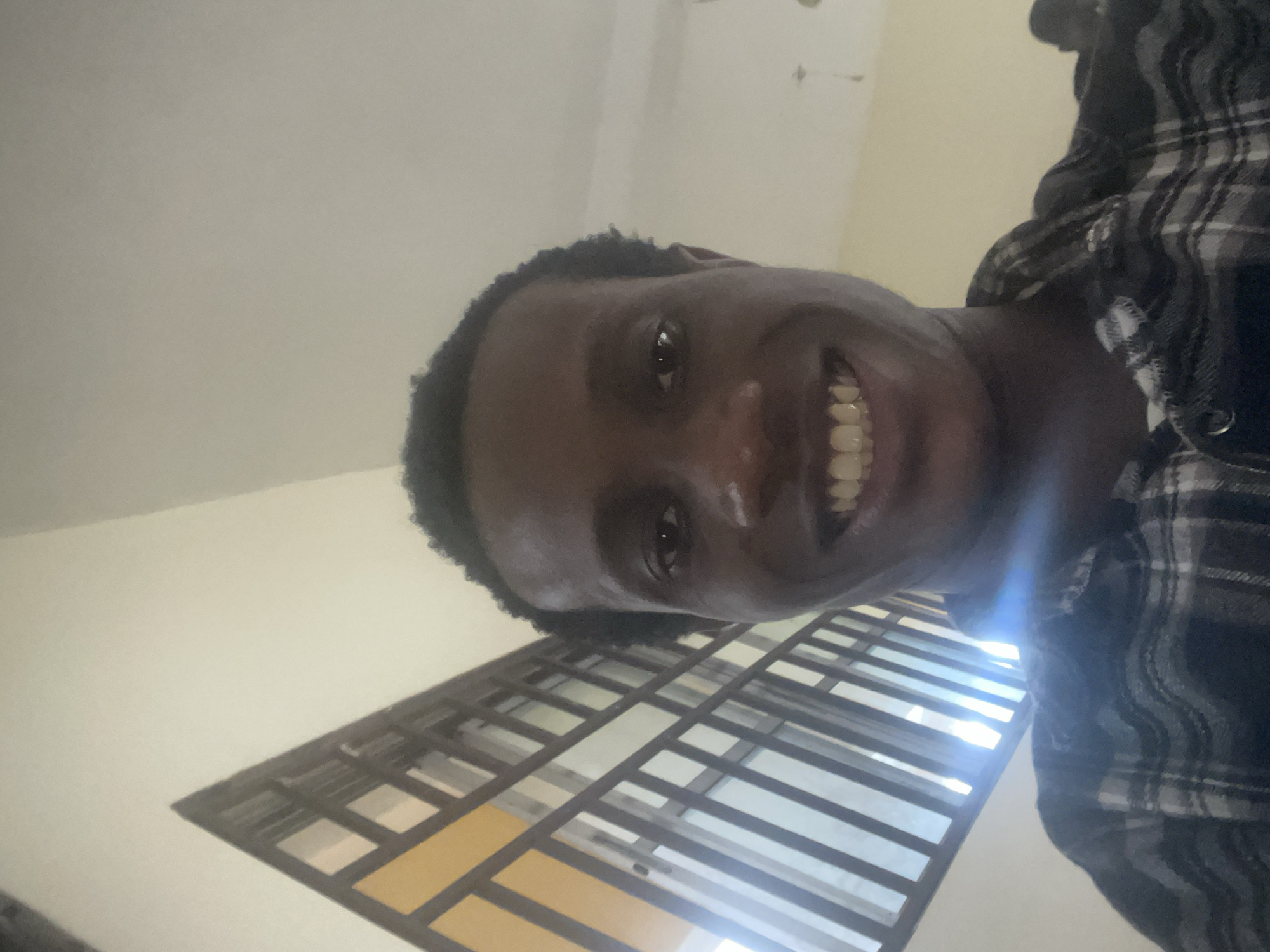 A close-up, low-angle selfie shows a young Black man with short, dark, curly hair, smiling widely with his teeth showing. He is wearing a black and gray plaid shirt. In the background, to the left, a triangular architectural feature with latticed windows is visible, and to the right, a light-colored wall. The lighting suggests an indoor environment, possibly a hallway or stairwell, with light streaming through the windows and creating some glare. The scene appears to be located in Ihiagwa, Nigeria.