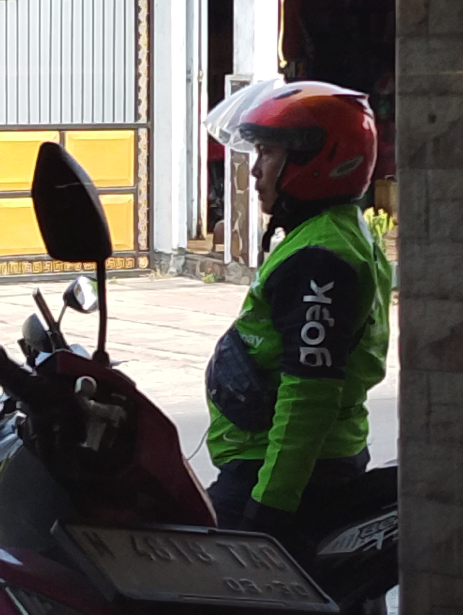 A person wearing a red helmet and a green jacket with "gojek" printed on the sleeve is sitting on a motorcycle. The person's face is not visible, but they appear to be looking to the right. The motorcycle has a black seat and a red front fairing. A license plate is visible on the front of the motorcycle, but the numbers are blurry. The background shows a building with yellow panels and a striped metal gate. The scene appears to be in an outdoor setting, possibly a street or a driveway. The image quality is low, with a blurry and pixelated appearance. The location of Manaruwi, Indonesia, is not evident from the image.