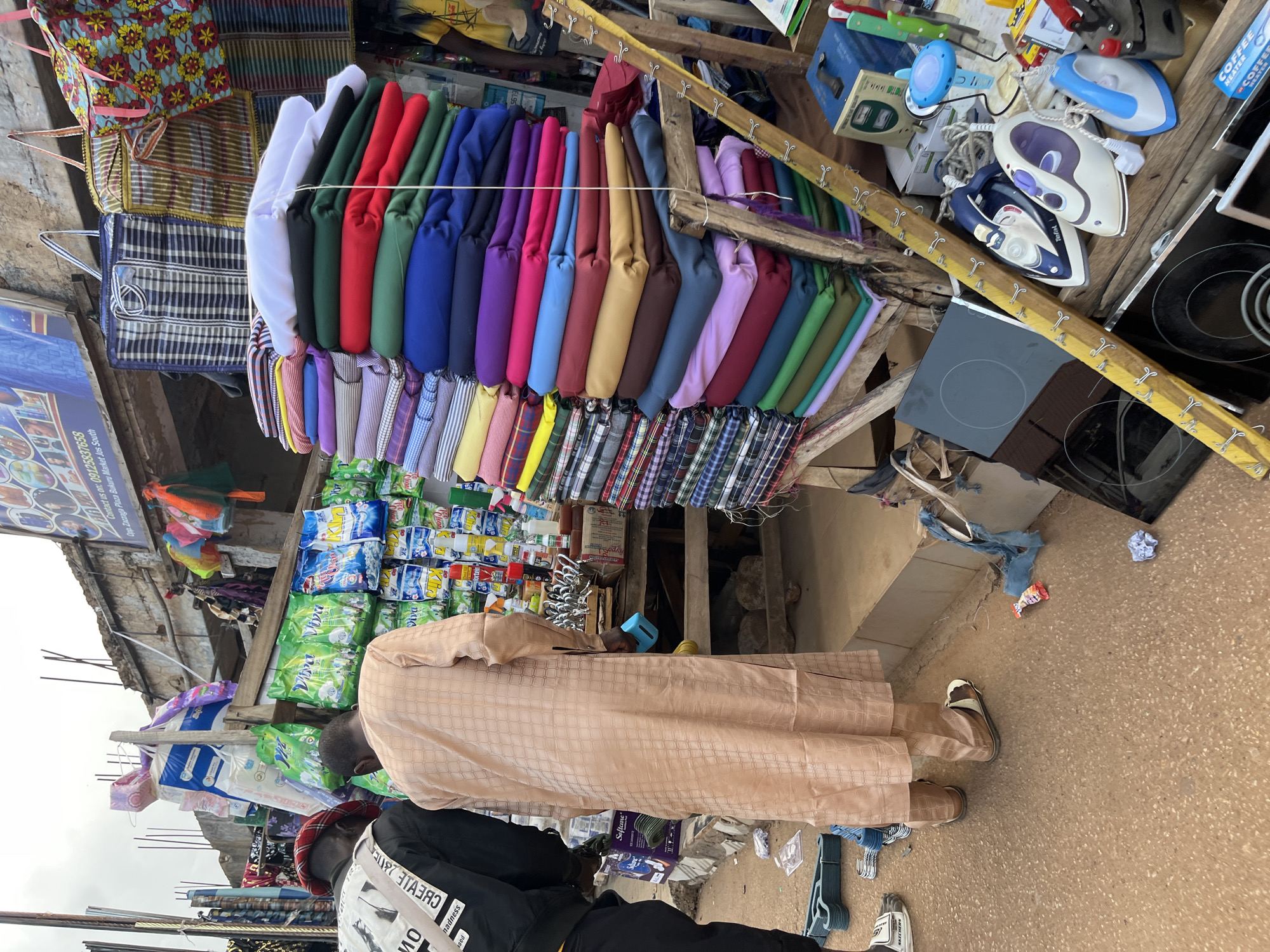 This image depicts an outdoor market scene in Bukuru, Nigeria. The foreground shows the lower bodies and feet of two individuals. The person on the left wears a black jacket with white lettering and a red beret. The person on the right is draped in a long, light brown garment with a checkered pattern and is holding a small blue object.
The market stall is laden with various items. Prominently displayed are stacks of colorful folded fabrics, neatly arranged by color. Among these fabrics, some appear to be patterned, while others are solid hues of red, blue, green, purple, and white. Adjacent to the fabrics are shelves filled with bags of cleaning products, featuring brands like "Viva." Further back, several large, woven bags with checkered and floral patterns hang from the stall's structure.
On a lower level of the stall, various household appliances are visible, including several electric irons of different designs and colors (white, blue, and black), and what appear to be induction hobs or stovetop elements. The stall itself is constructed from rough wooden planks and salvaged materials.
The background reveals the weathered facade of a building with exposed rebar, suggesting ongoing construction or repair work. A banner above the stall displays text, including a phone number and the location information "Opp. Zazaga Plaza Bukuru Mkt. Jos South." The ground is a rough, reddish-brown textured surface, possibly asphalt or compacted earth, with scattered debris, including plastic wrappers and a crumpled piece of paper. The overall impression is a bustling, functional market environment.